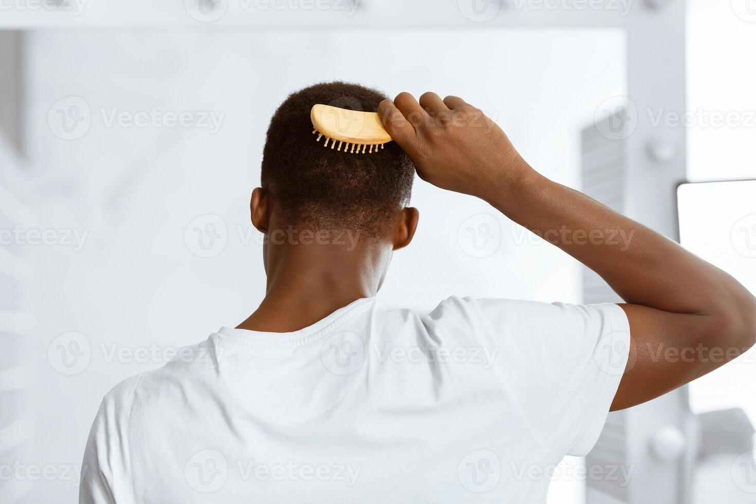 African American Guy Brushing And Combing Hair With Hairbrush Standing Back To Camera In Modern Bathroom At Home. Male Haircare, Men's Daily Beauty Routine Concept. Rear View photo