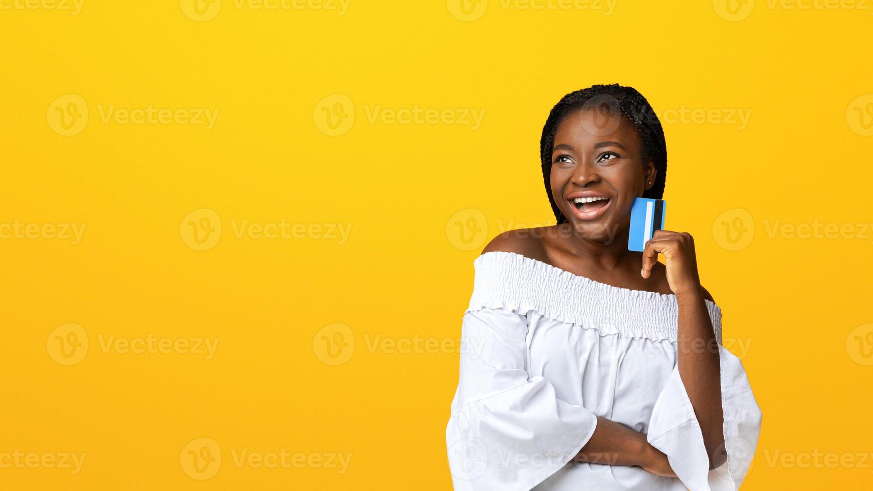 Positive black lady dreaming about shopping with credit card in hands, african american shopaholic lady looking away with pensive face expression, isolated over yellow background, copy space photo