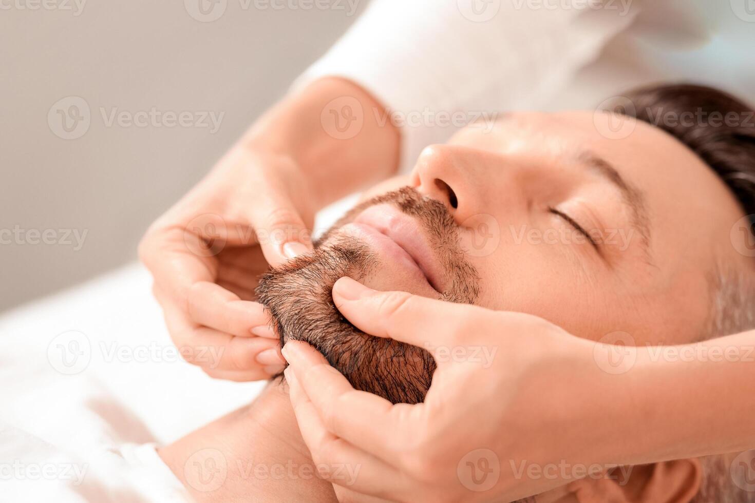 Closeup of middle aged man getting healing face massage at luxury spa. Spa therapist making relaxing massage for handsome man, rubbing his bearded chin, copy space. Male cosmetology concept photo