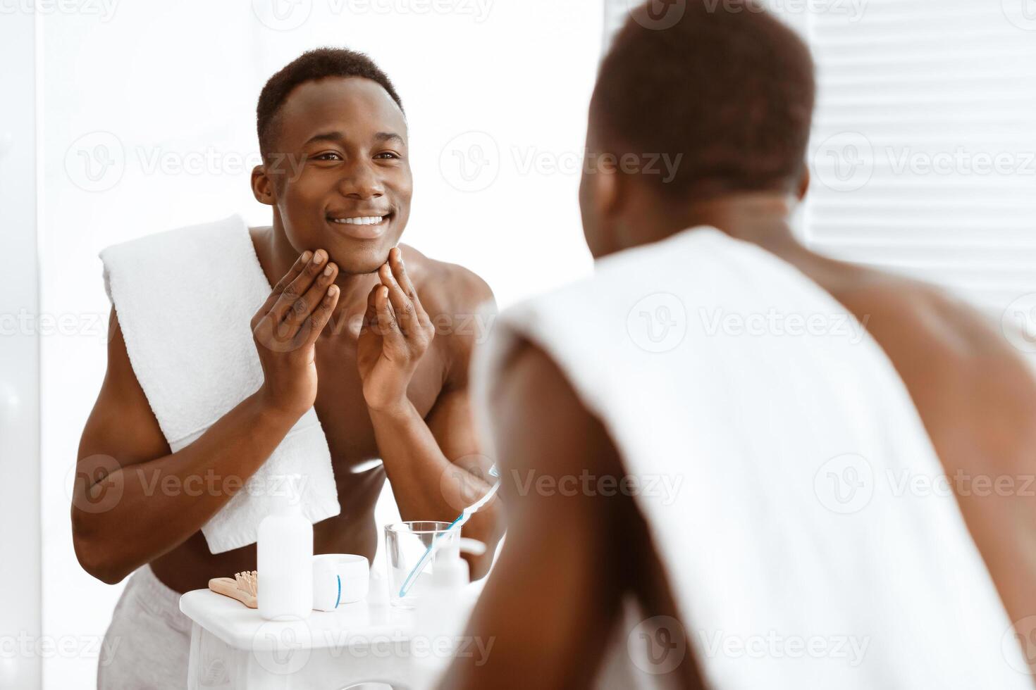 Happy Black Man Touching Smooth Face Skin After Shaving Standing Near Mirror In Bathroom At Home. Male Facial Skincare, Morning Beauty Routine And Self-Care Cosmetics Concept photo