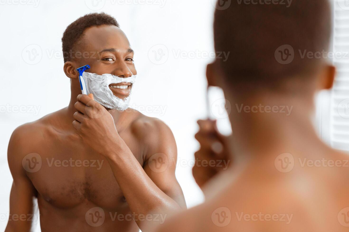Cheerful Shirtless Black Guy Shaving Face Using Razor And Shave Foam Standing Near Mirror In Modern Bathroom At Home. Male Facial Skincare And Morning Beauty Routine Concept photo