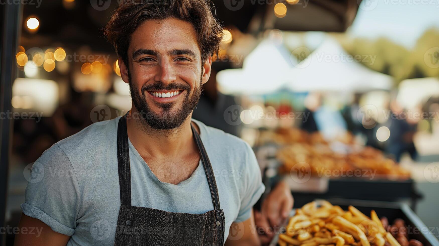 A young man with a friendly smile stands behind a food stall offering crispy fries. The lively outdoor market buzzes with activity in the background under the warm afternoon sun. photo