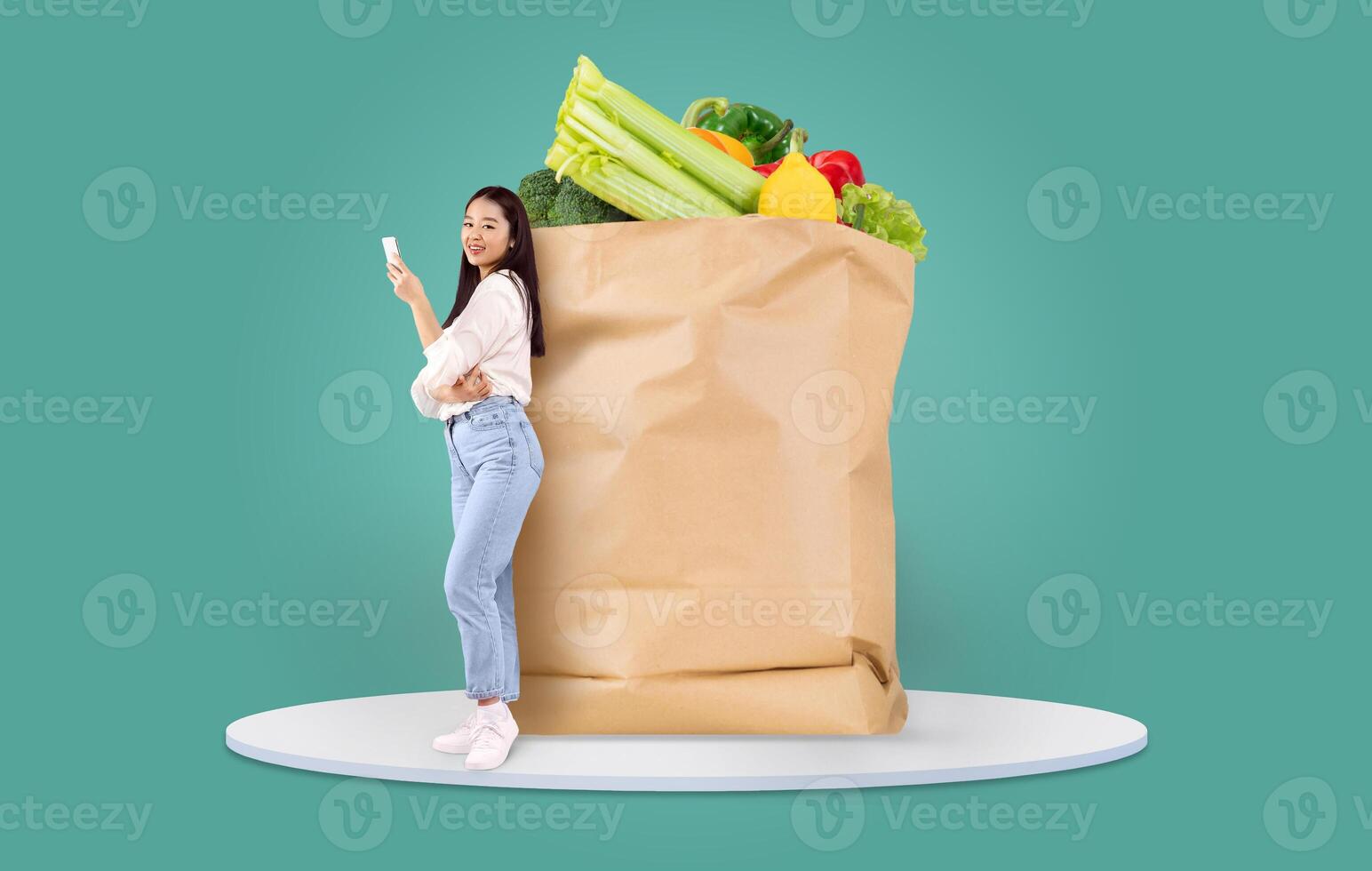 A customer stands next to a large paper bag filled with fresh groceries, using a mobile device to manage her shopping. Online grocery shopping has become a popular choice during recent times. photo