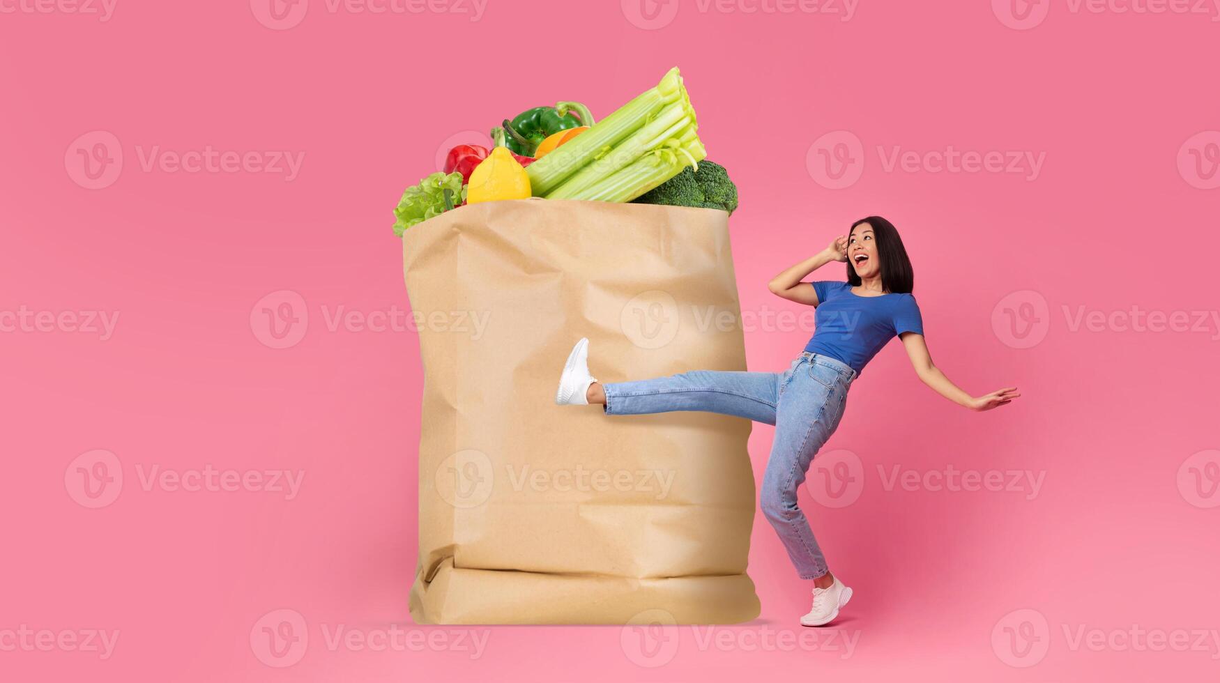 A cheerful customer stands beside a large paper bag filled with colorful groceries. The background is vibrant pink, highlighting the joy of shopping for fresh food essentials in a modern lifestyle. photo