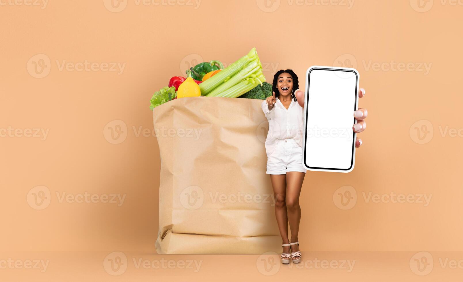 A customer stands beside a large paper bag filled with fresh vegetables and fruits while holding a smartphone. This reflects the modern shopping experience in grocery stores. photo