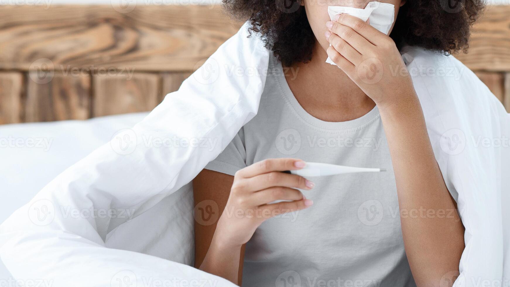 Home treatment from flu and colds, viral diseases and feeling unwell. Desperate young african american female sits on bed, wiping nose, looks in thermometer at interior of bedroom, free space, cropped photo