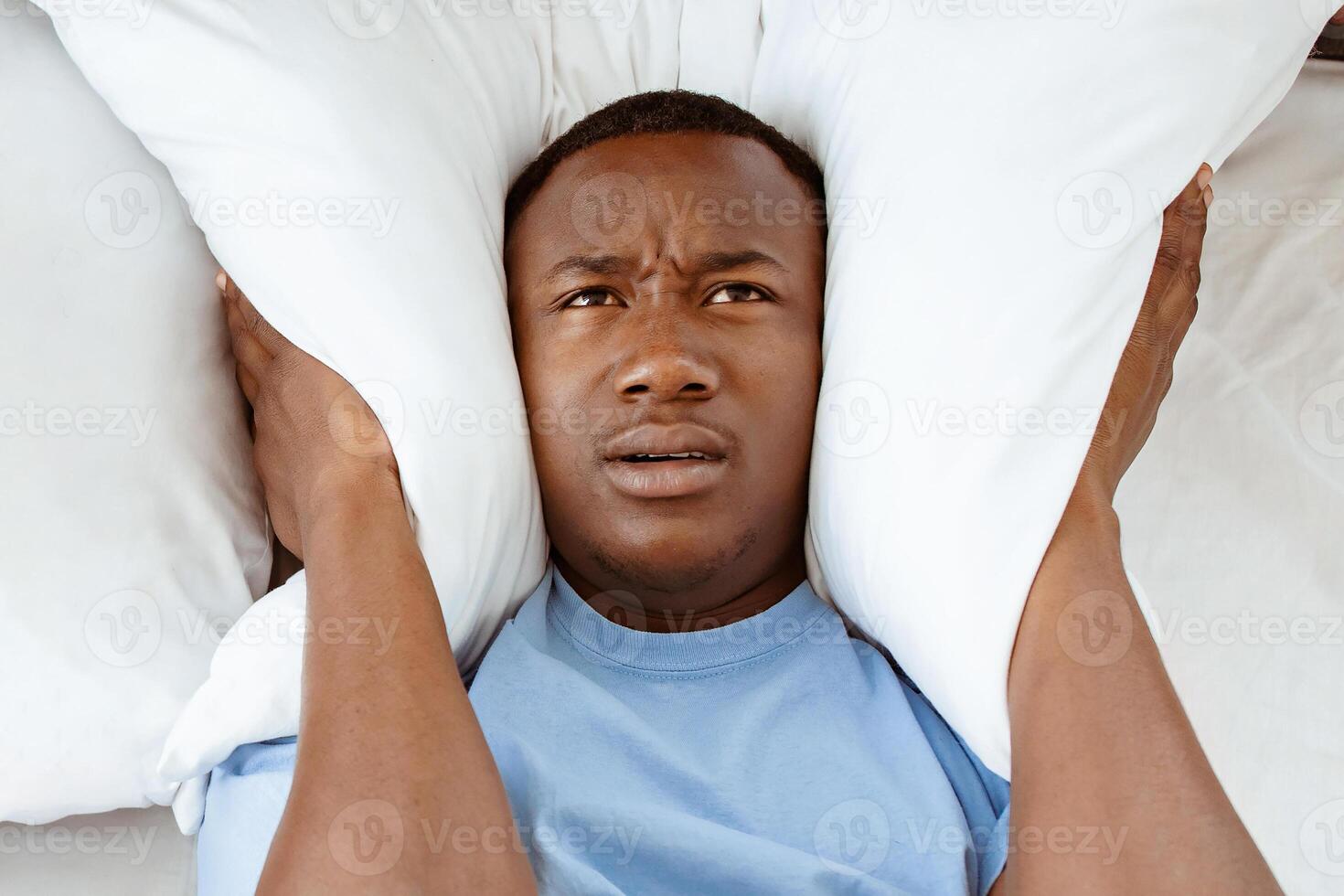 Snoring, insomnia, bad sleep, loud neighbors. Upset tired young african american man freaking out, covers his ears with pillow so as not to hear noise in bedroom, vertical, top view, copy space photo