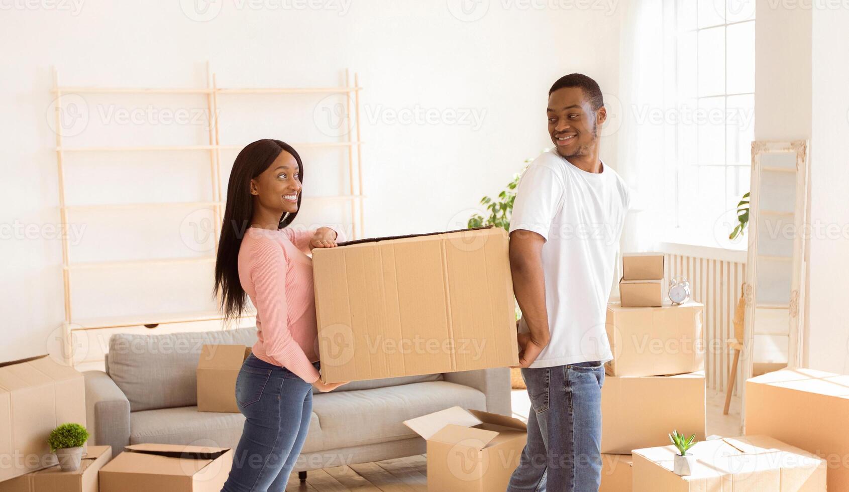 Loving black couple carrying cardboard box together, helping each other in their new home on moving day. African American tenants with belongings relocating to new apartment, panorama photo