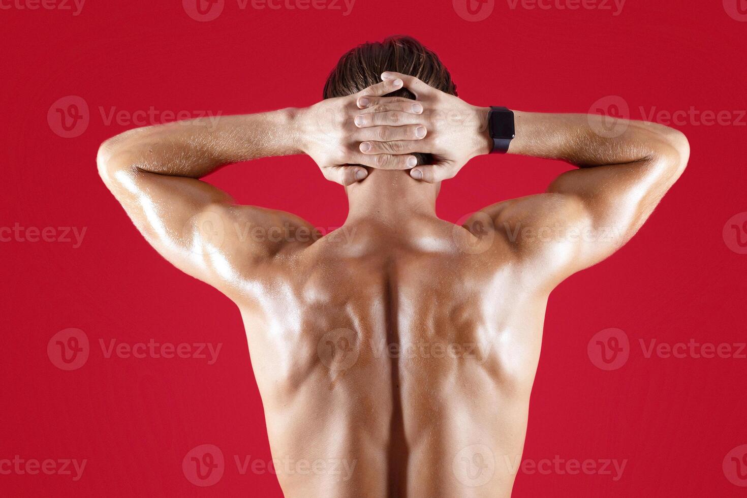Handsome young sportsman with bare back standing with hands behind his head on red studio background. Shirtless millennial guy with sexy body posing and demonstrating his muscles photo