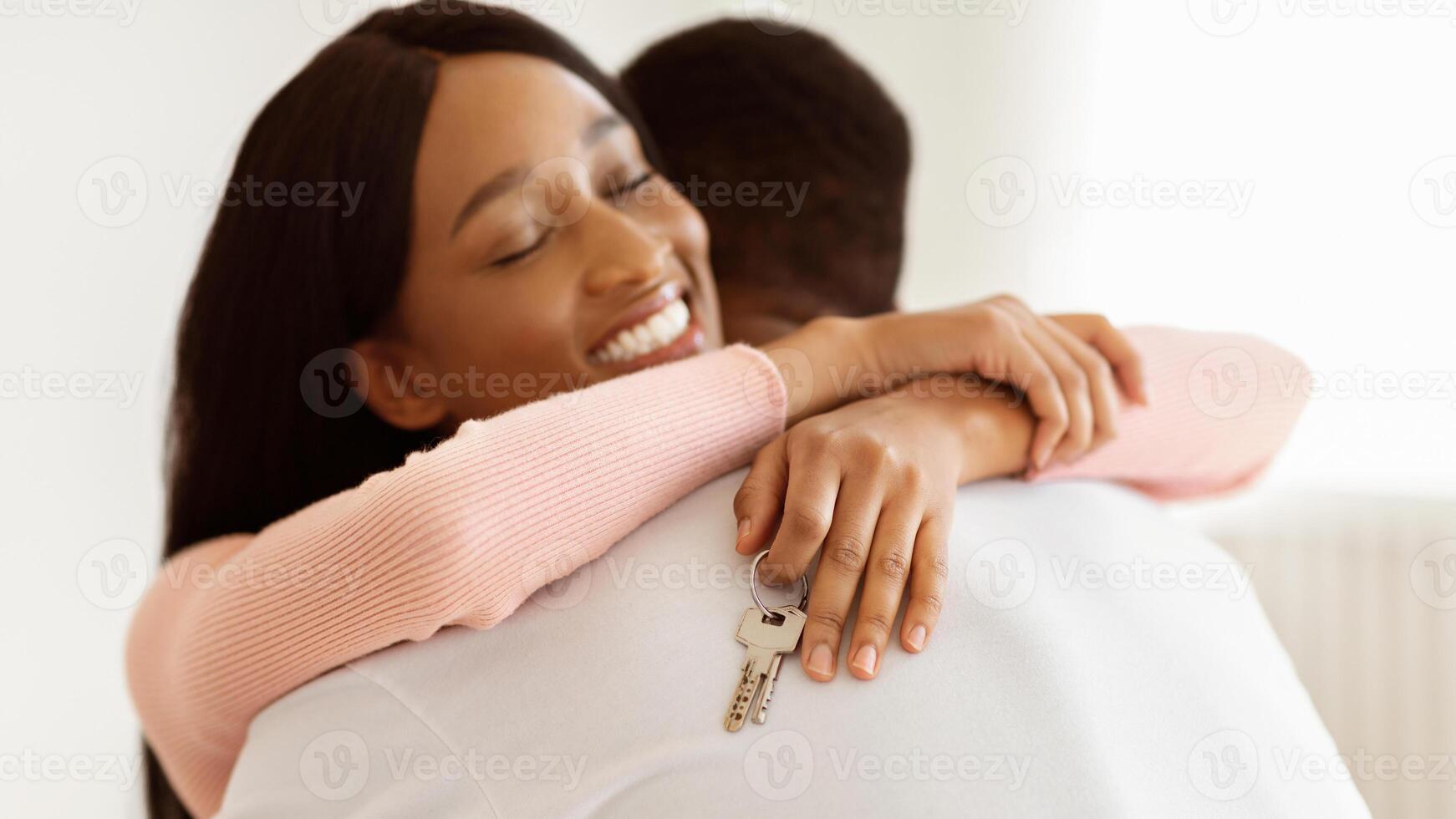 Smiling black woman holding house key and hugging her husband indoors. Millennial African American lady happy over moving to new apartment with her boyfriend. Panorama photo