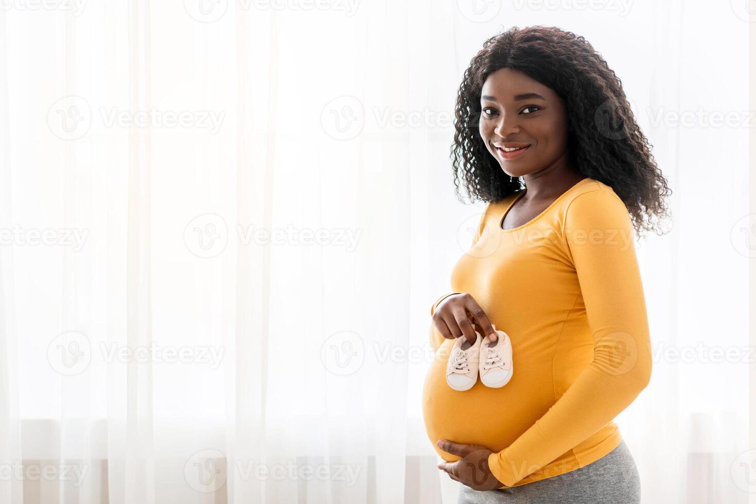 Motherhood concept. Happy african american expecting woman showing small shoes for her coming baby, copy space. Pregnant black woman holding baby shoes on her belly, standing by window at home photo