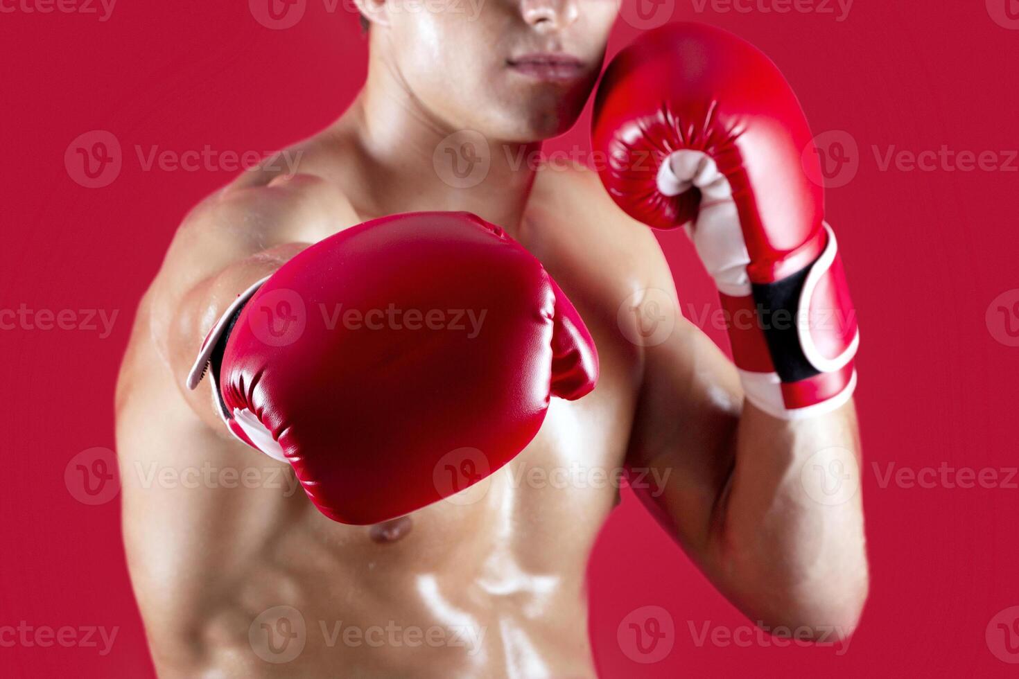 Determined young boxer in fighting gloves making punch at camera on red studio background, selective focus. Unrecognizable millennial sportsman with bare chest fighting, cropped view photo