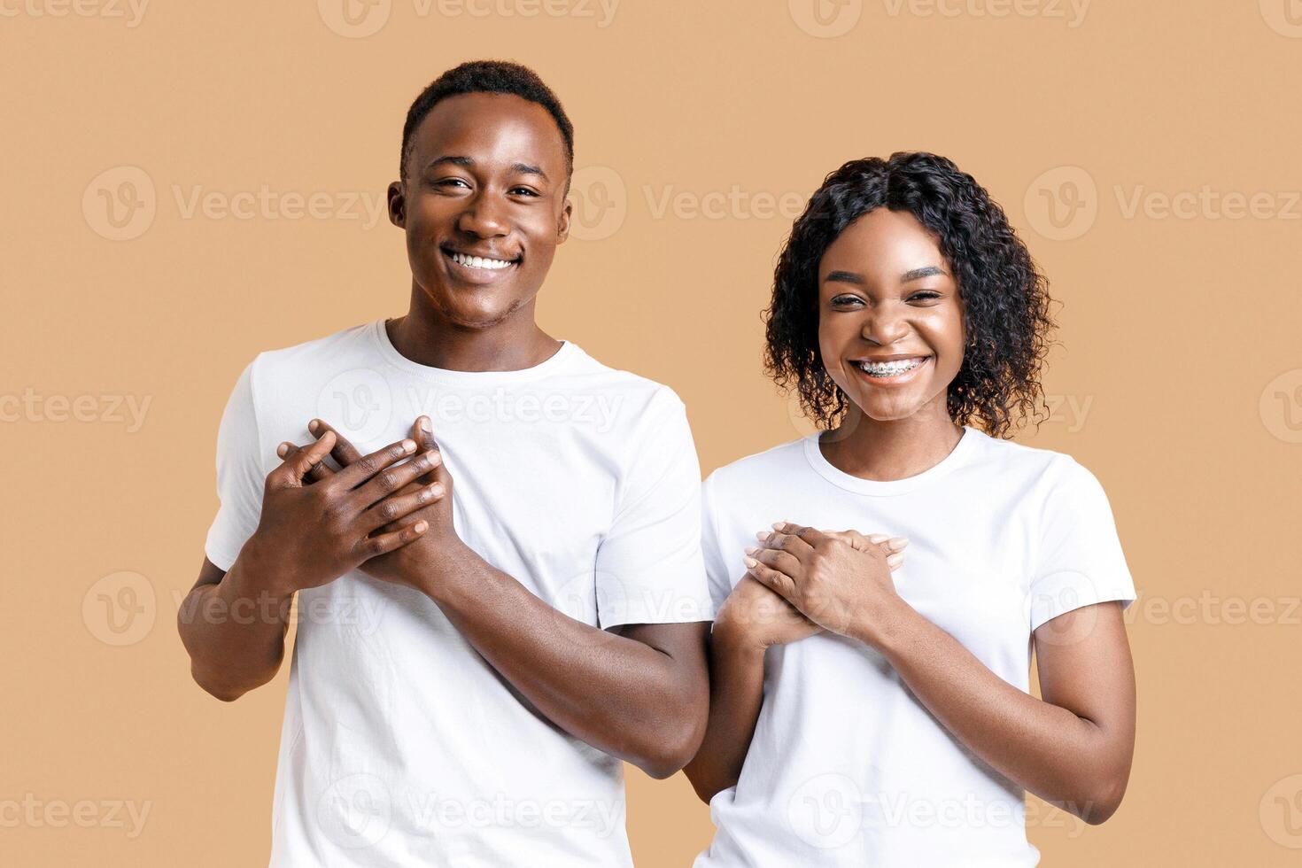 Grateful african-american couple keeping both palms on chest and smiling, expressing gratitude and good feelings, yellow studio background. Happy black man and woman hoping for better photo