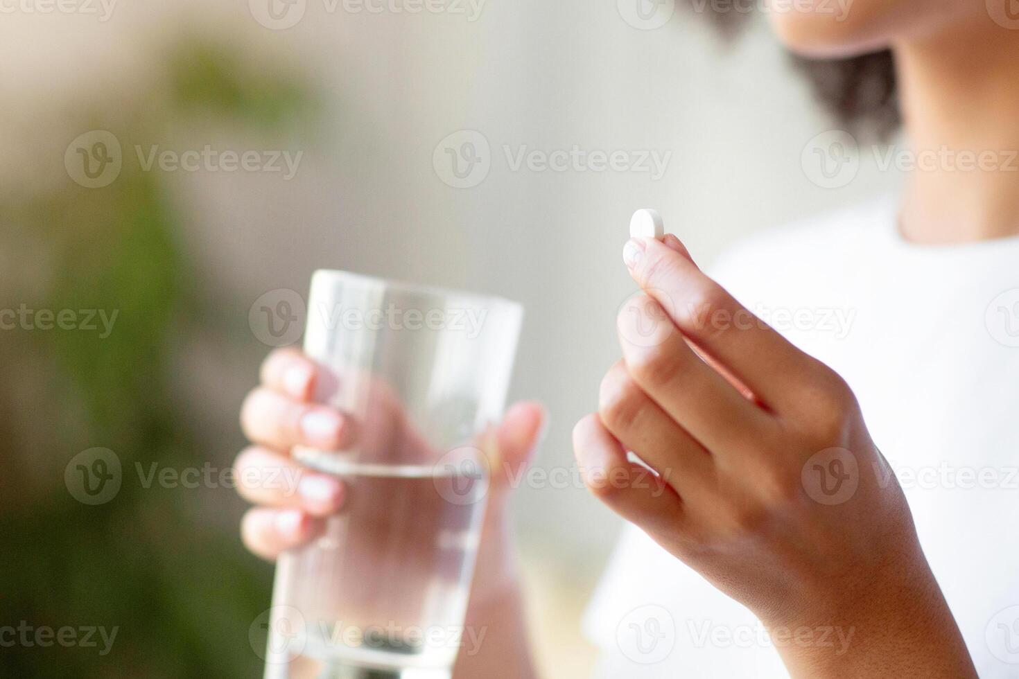 Smiling woman taking white round pill, holding water glass in hand. Happy young female taking supplement, daily vitamins for hair, skin, body, natural beauty and healthy lifestyle, free space, cropped photo