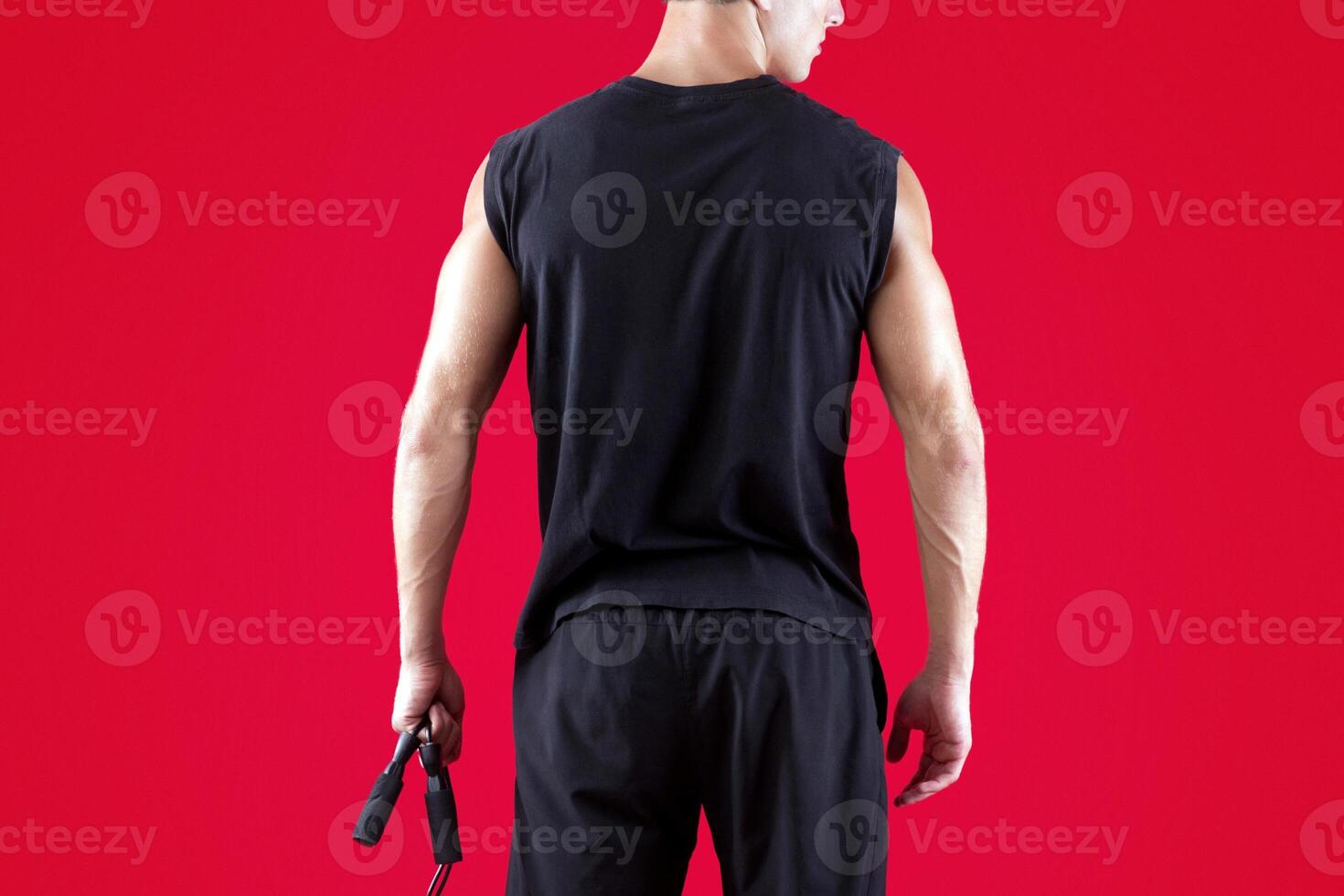 Aerobic workout. Back view of strong young man standing with skipping rope on red studio background. Full length portrait of motivated athlete warming up before training, doing cardio exercises photo