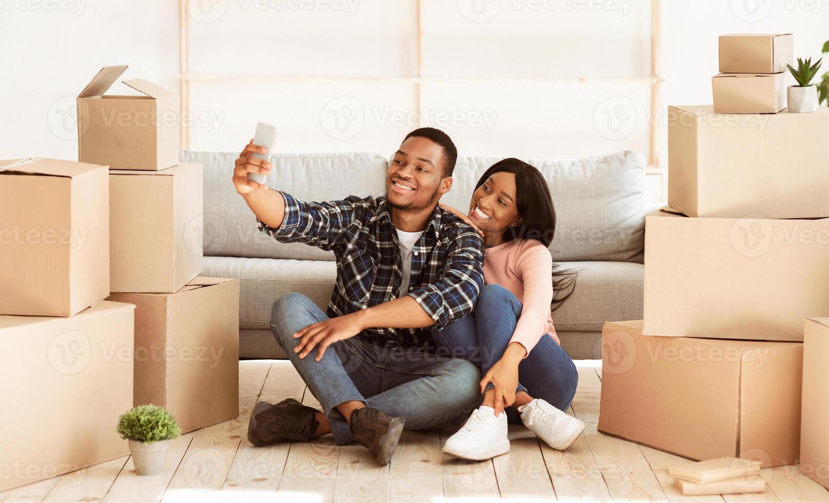 Smiling black guy and his girlfriend moving to their own home together, taking selfie among carton boxes, panorama. Happy African American couple making photo of first day in new apartment