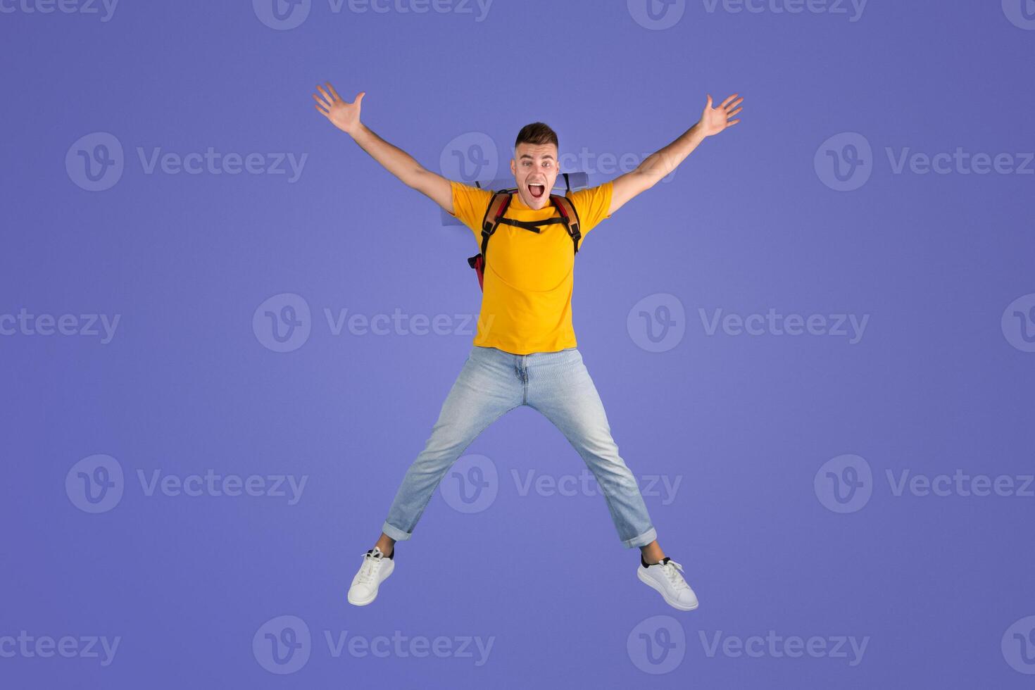 Handsome millennial guy with backpack jumping in excitement over blue studio background. Young male traveler happy to go on hiking journey or camping trip. Tourism concept photo