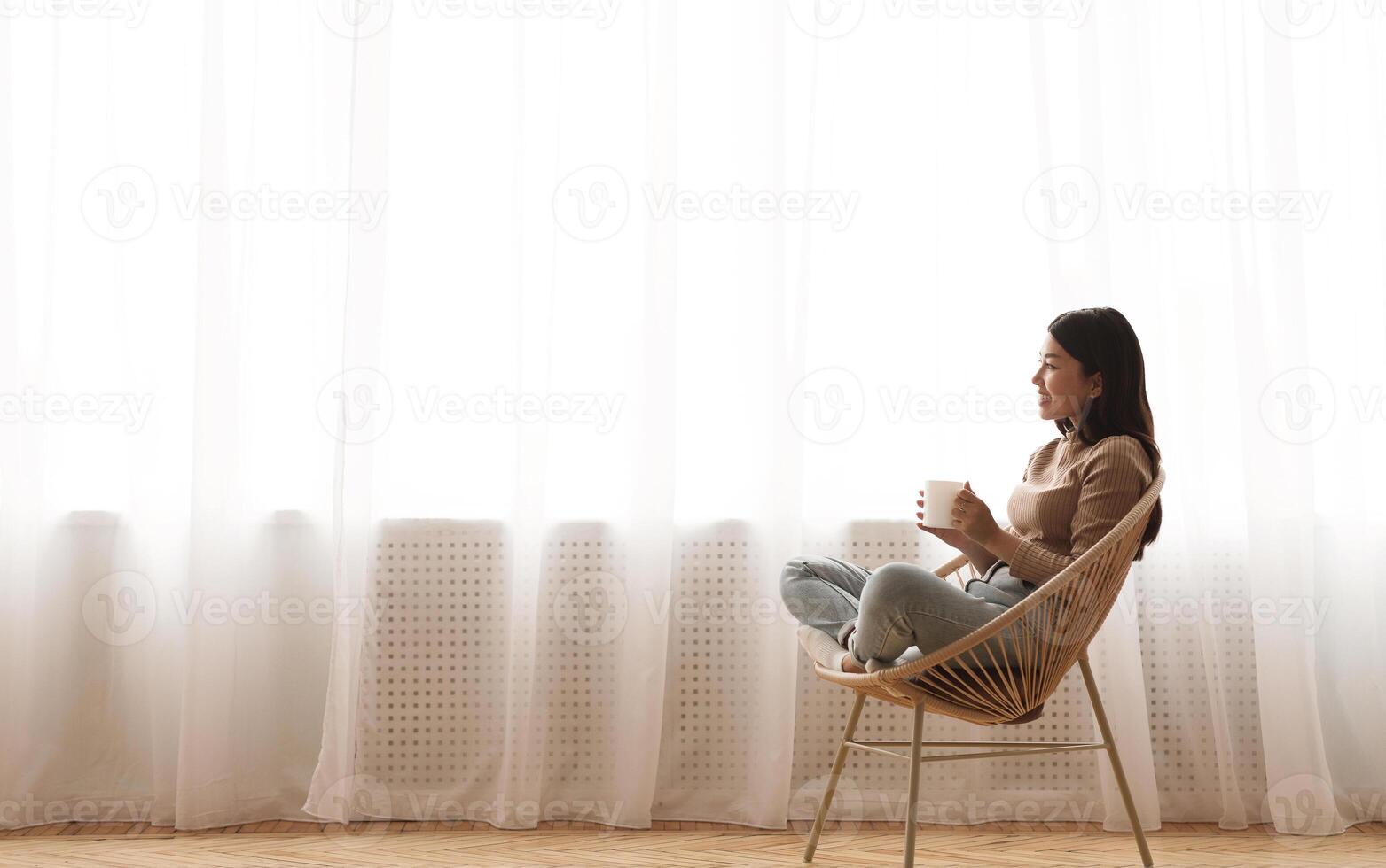 Tea time. Relaxed girl sitting in modern chair, enjoying hot coffee in front of window, side view, free space photo