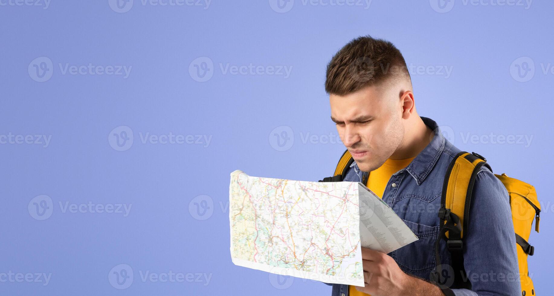 Puzzled young guy with tourist backpack and map over blue studio background. Funky male backpacker having difficulty planning his travel route, lost in big city photo