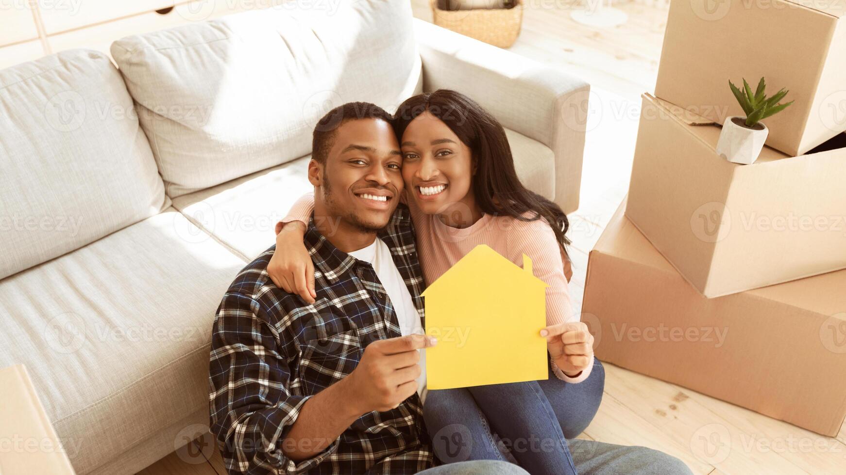 Black millennial guy with his girlfriend hugging and holding paper house while sitting on floor of their new apartment on moving day. Relocation, tenancy, home rentals concept photo