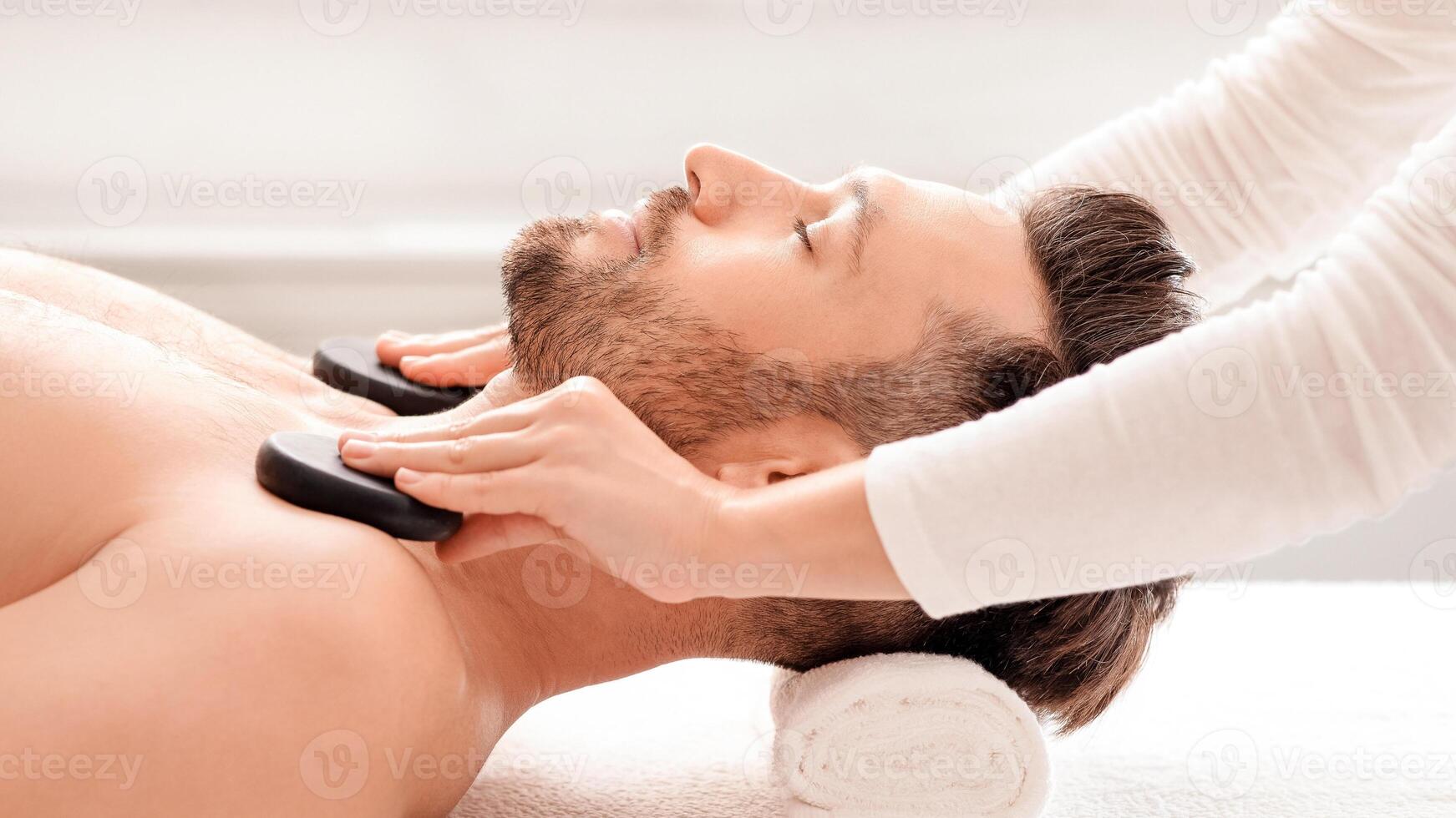 Thai spa for men. Unrecognizable female therapist placing black stones on relaxed man shoulders, side view. Middle-aged wealthy man enjoying day at male spa, having healing hot stone massage photo