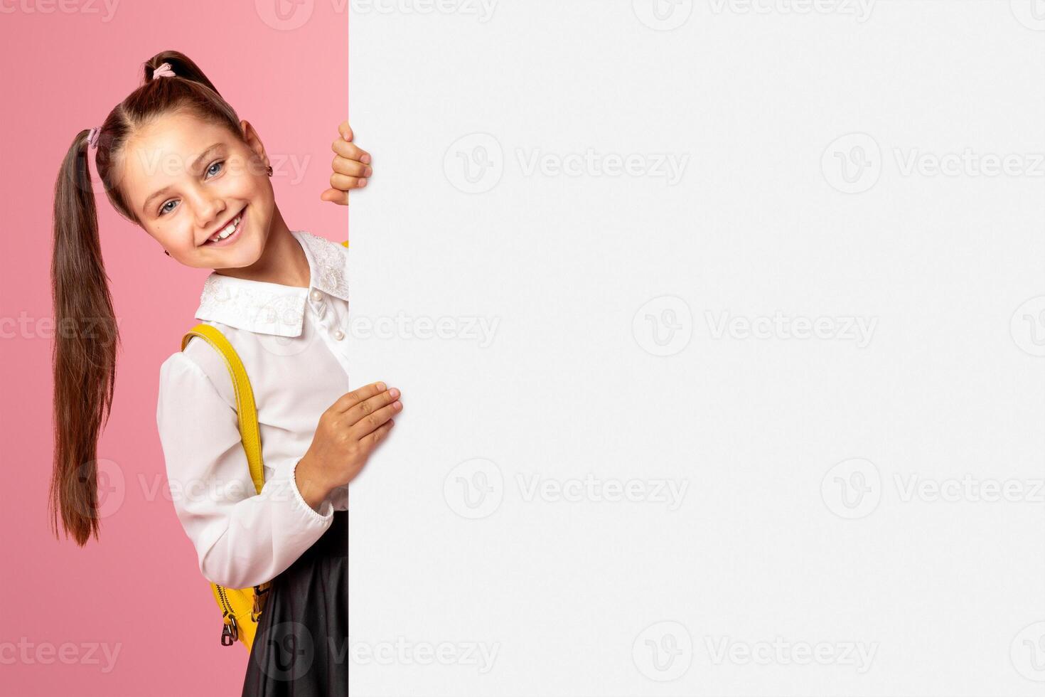 Ready for lesson. Cheerful schoolgirl in uniform peeps out from with white billboard with copy space, studio shoot photo