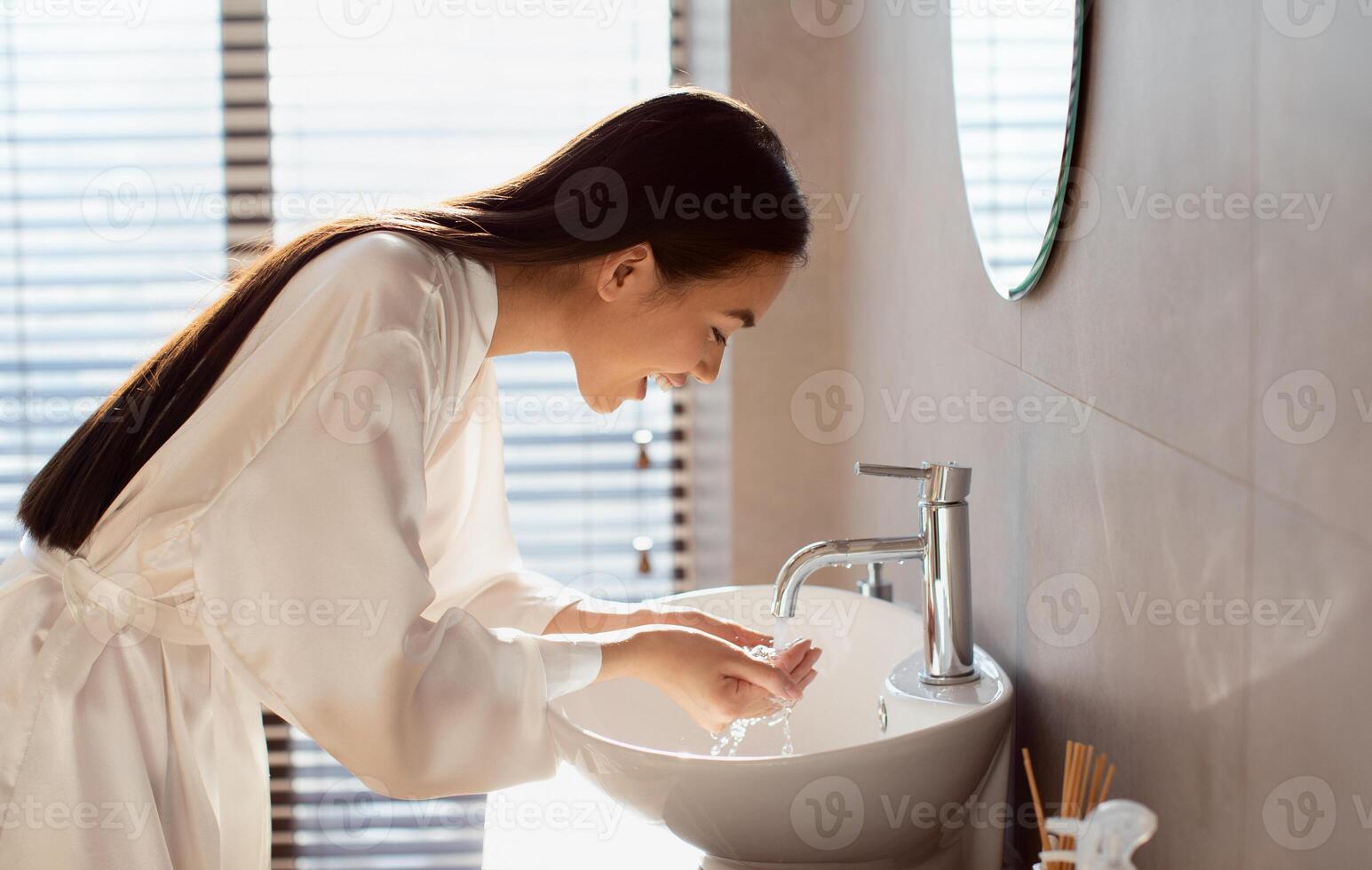 Pretty long-haired young japanese lady in silk bathrobe washing face in the morning after waking up, beautiful bathroom interior, side view, copy space. Hygiene, body care, skin care concept photo