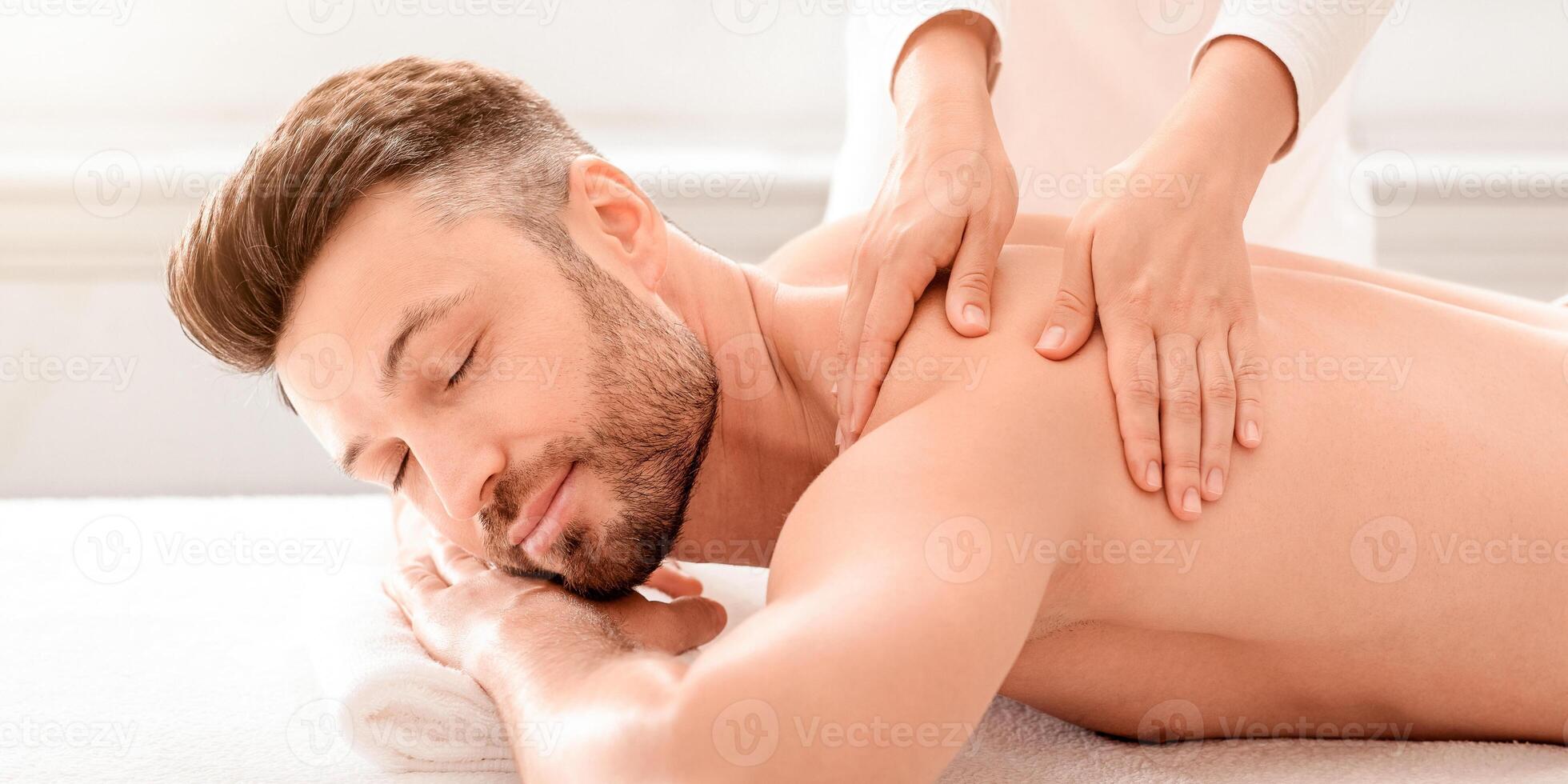 Unrecognizable female therapist rubbing joyful man shoulders at modern spa salon, side view. Handsome bearded middle-aged man relaxing while getting healing back massage photo