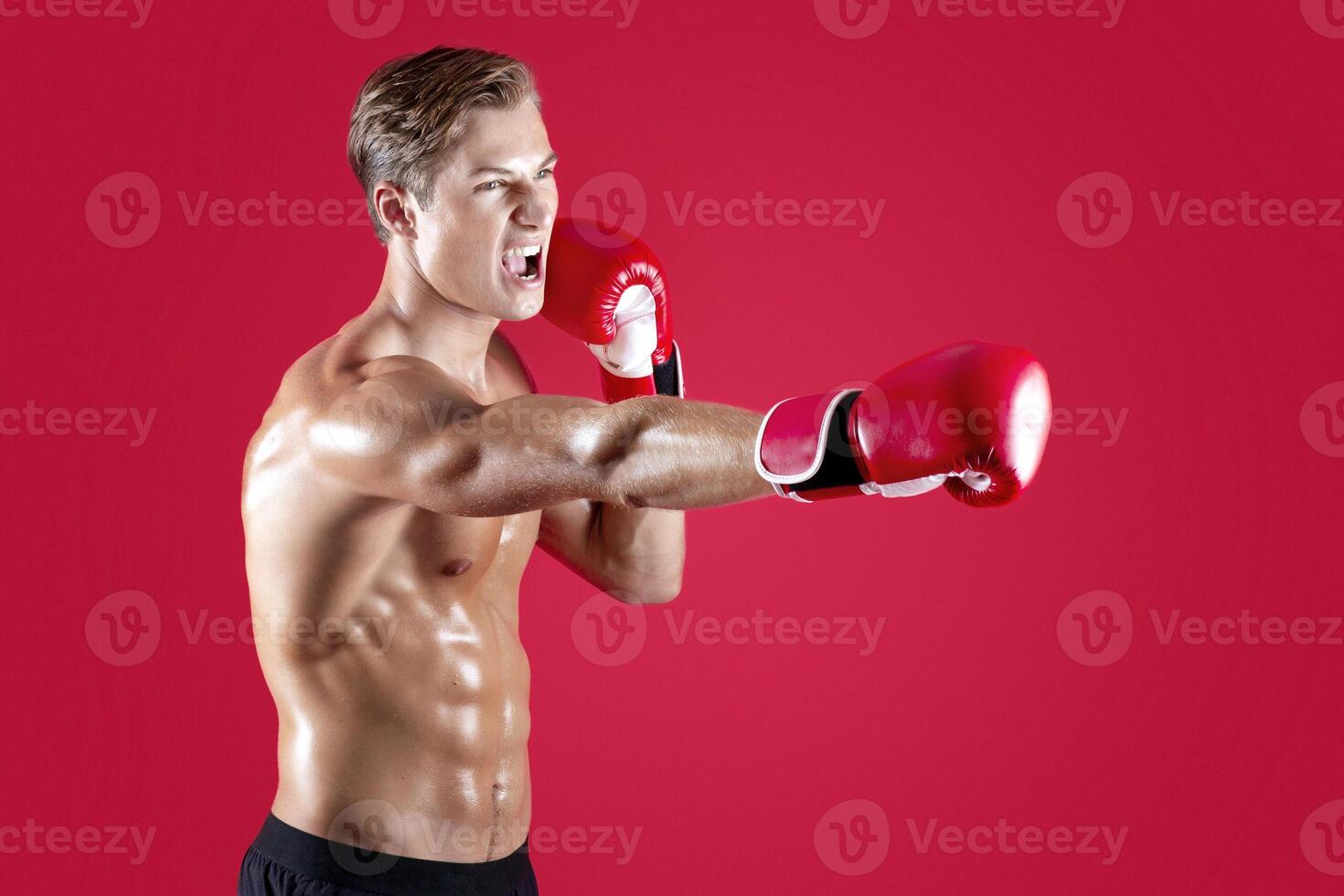 Powerful millennial athlete in boxing gloves shouting in anger and making punch on red studio background,copy space. Young MMA fighter with sexy bare torso fighting and screaming photo