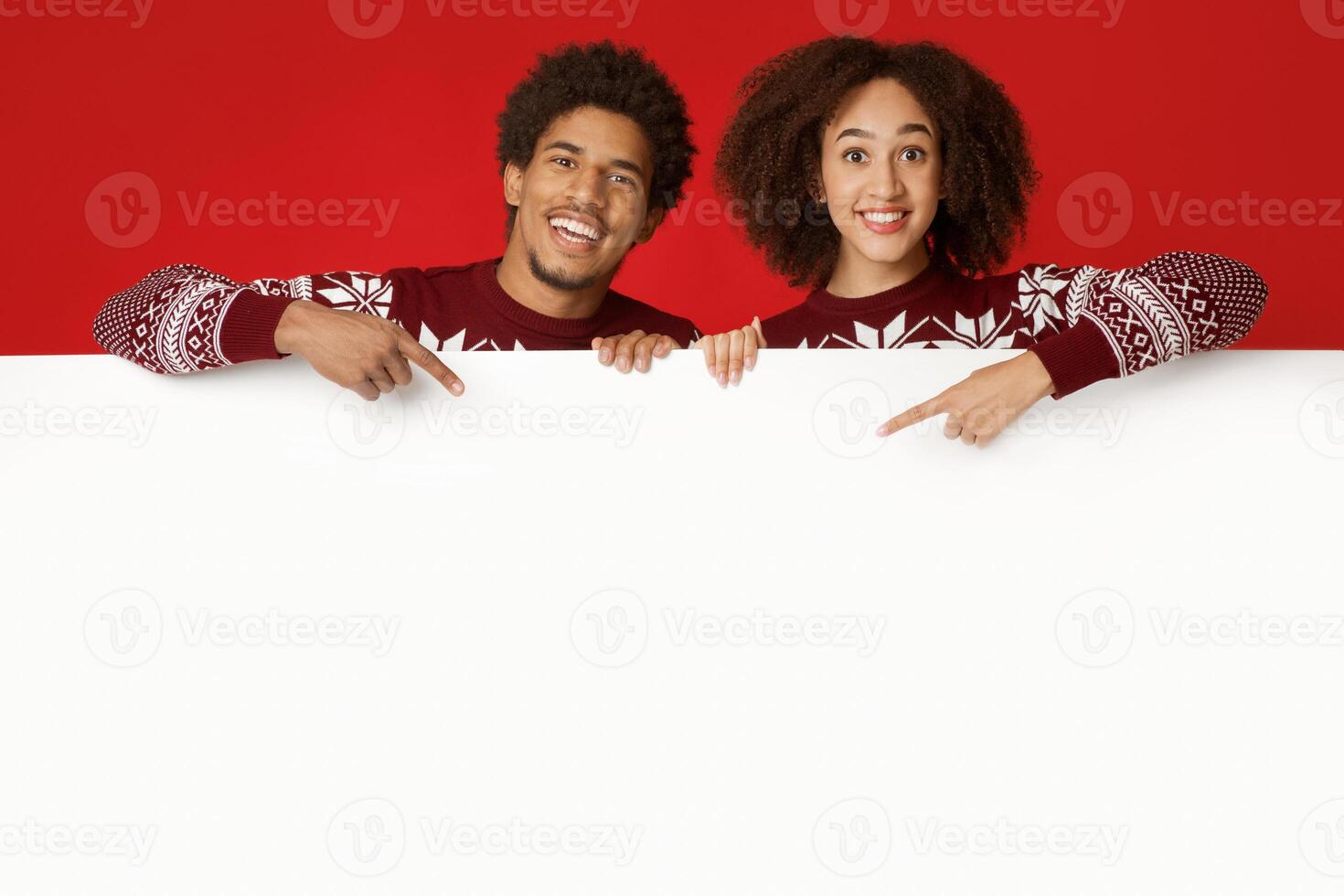 Happy New Year and winter holidays celebration. Smiling millennial african american lady and man in same Xmas sweaters holds white billboard and points at it with fingers, isolated on red background photo