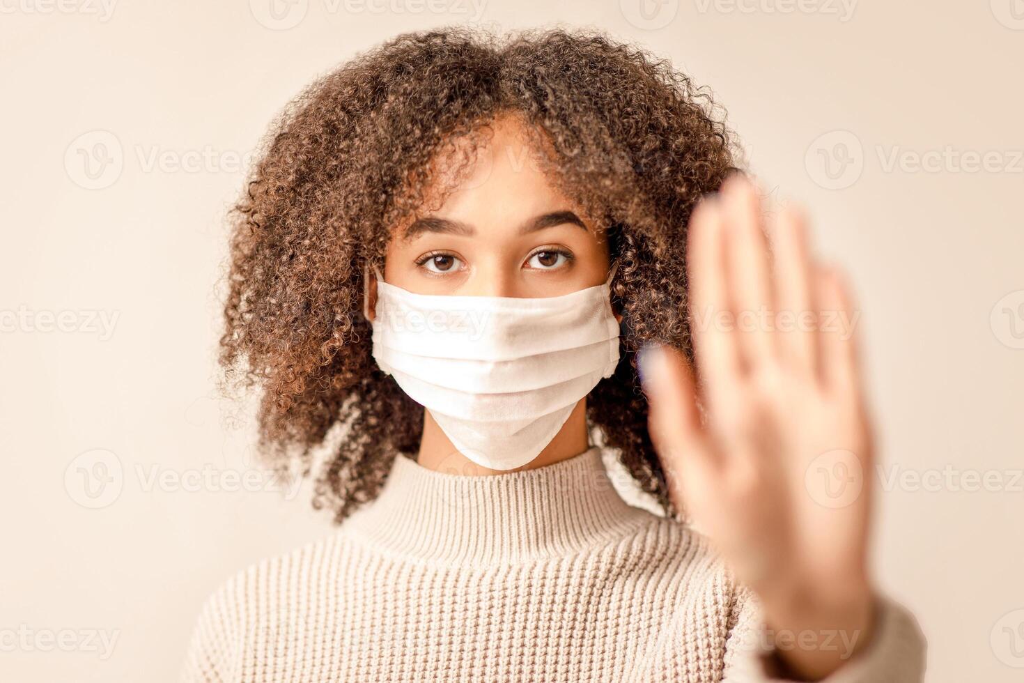 Stop coronavirus and health protection during epidemic. Serious african american young female in protective mask making hand gesture, focus on face of lady in sweater isolated on light background photo