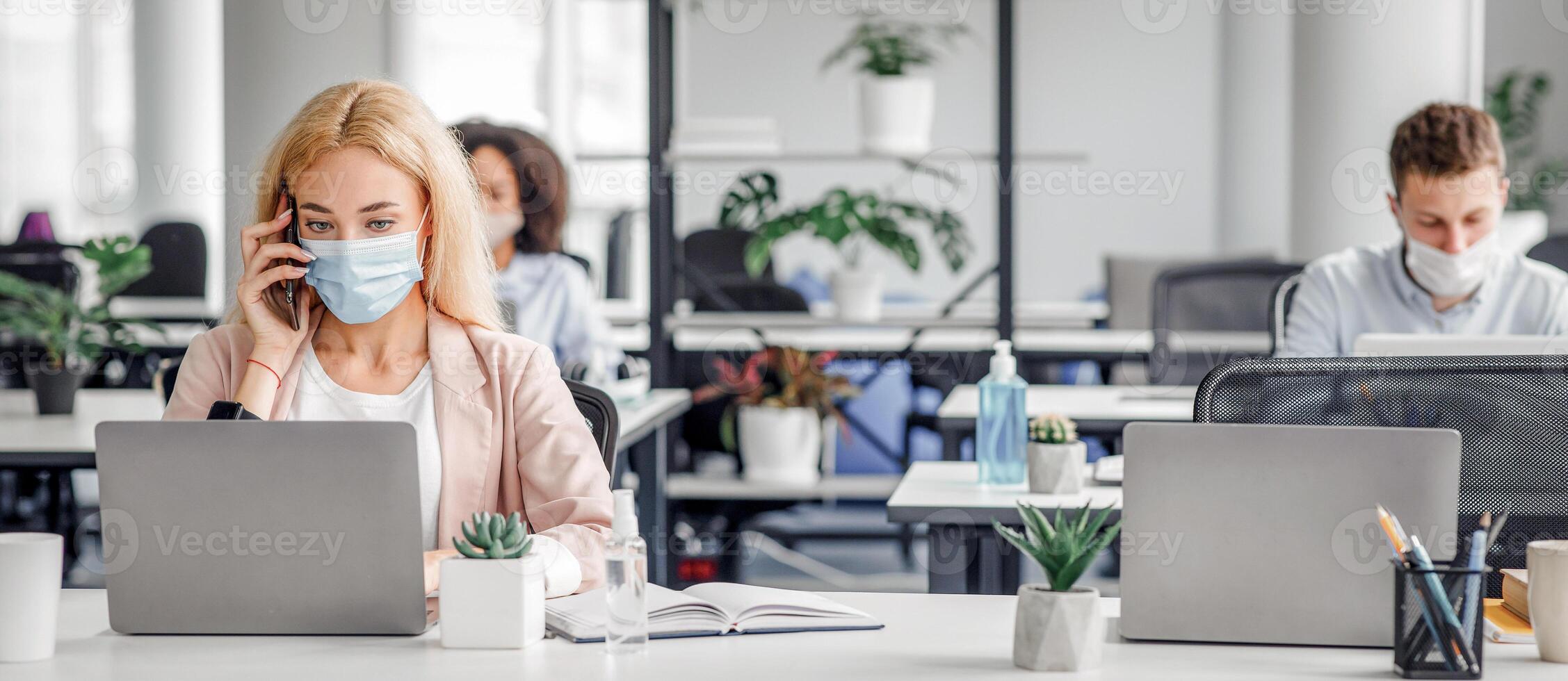 Working with client by phone remotely during COVID-19 outbreak. Millennial woman in protective mask talking at smartphone and typing on laptop in modern office interior, copy space photo
