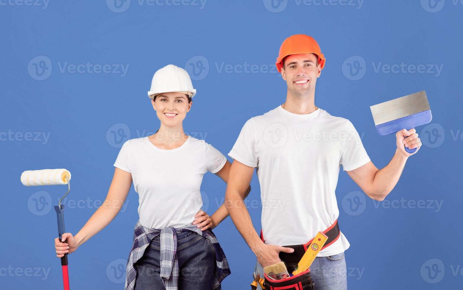 Renovation Time. Positive Couple Posing With Putty Knife And Roller Painter Over Blue Studio Background, Happy Confident Man And Woman In Hard Hats Ready For Repair Works, Copy Space photo