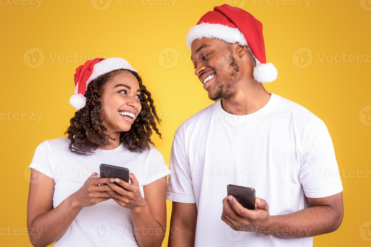 African American Couple In Santa Hats Using Smartphones Celebrating Christmas, Smiling Each Other To Camera Standing Over Yellow Studio Background. Mobile Application Xmas Advertisement. Panorama photo