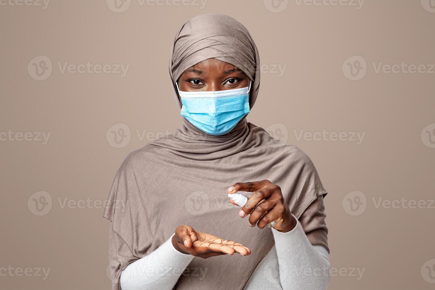 Coronavirus Precautions. Black muslim woman in medical mask applying disinfectant spray on hands to prevent germs spreading, islamic lady in hijab posing over light studio background, copy space photo