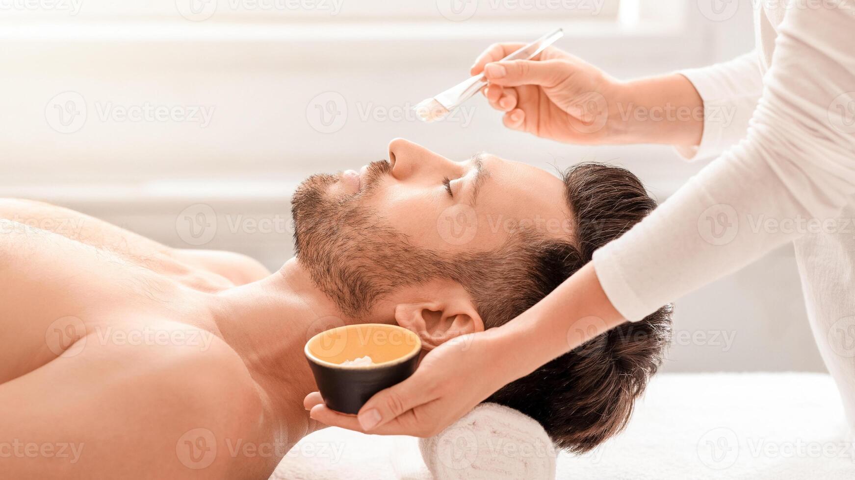 Handsome bearded man having face treatment at male spa salon, side view, copy space. Relaxed middle aged man enjoying day at luxury spa, getting anti-aging face mask, closeup photo