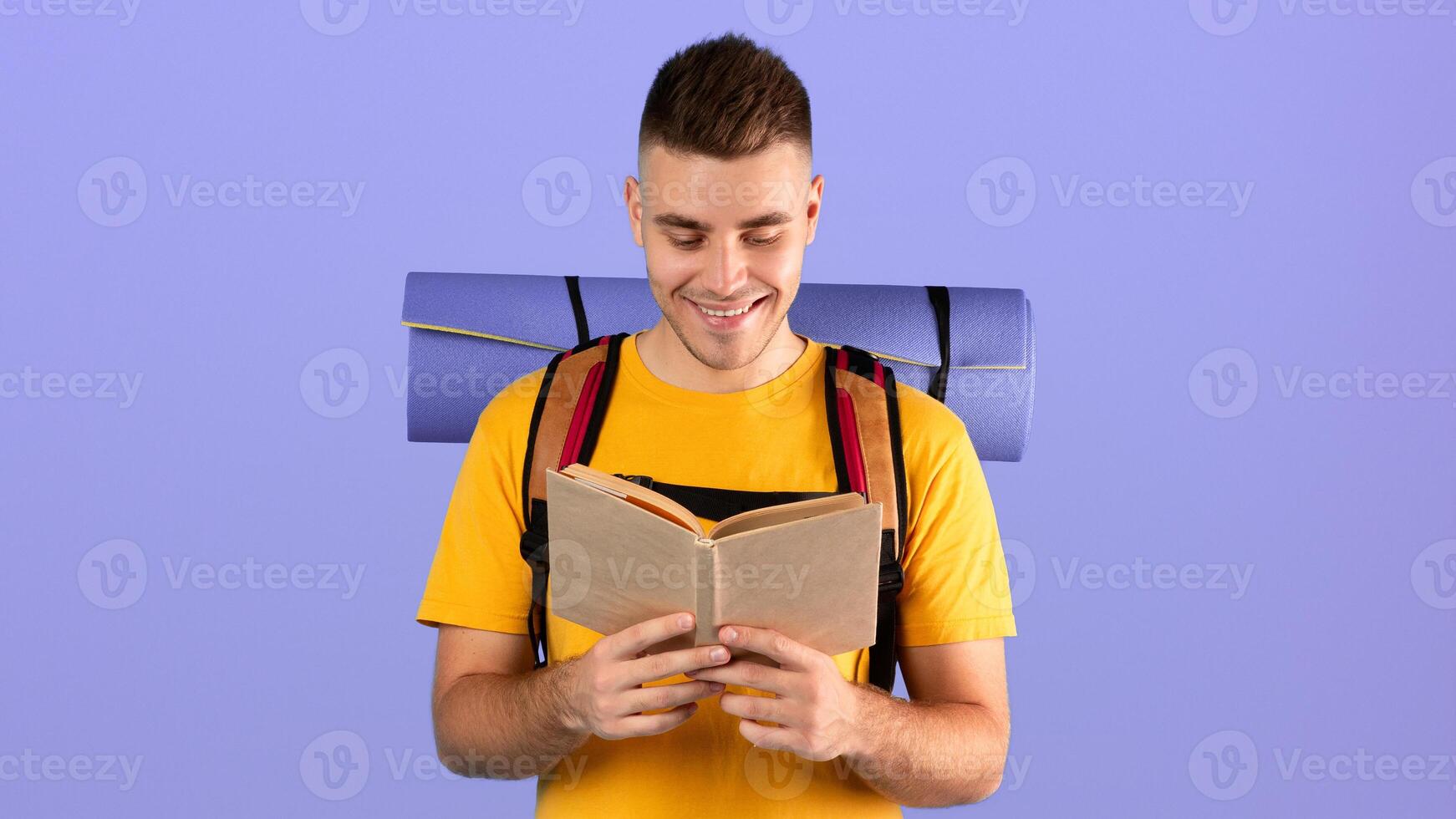 Positive millennial hipster with camping gear reading dictionary or guidebook over blue studio background. Happy young guy looking through conversation book while traveling abroad photo