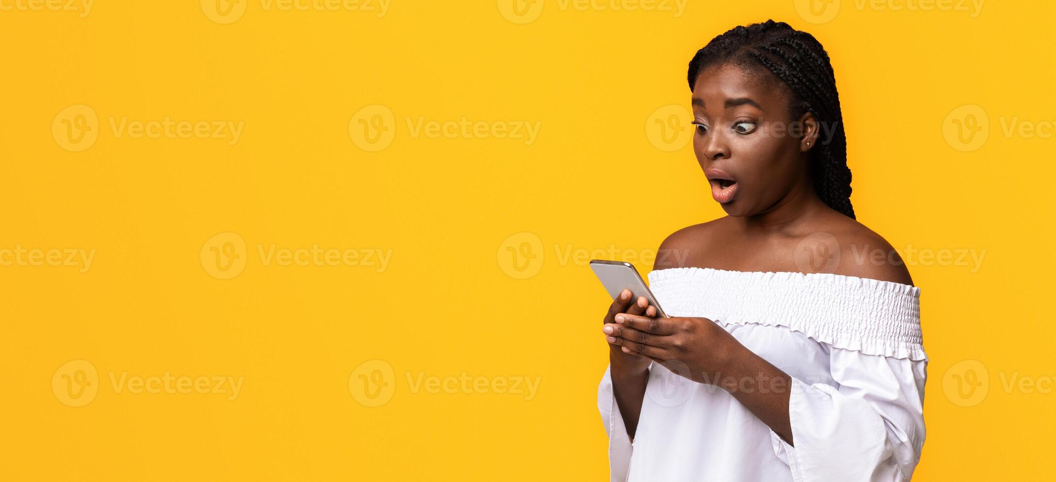 Big Online Sales. Shocked African American Woman Holding Smartphone, Looking At Device's Screen With Amazement, Emotionally Reacting To Mobile Offer, Standing On Yellow Studio Background, Copy Space photo