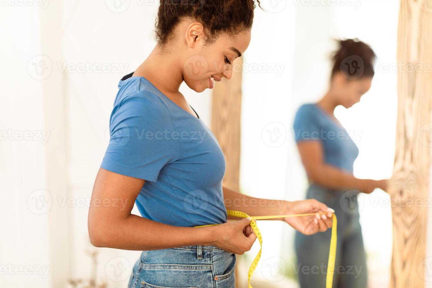 Smiling African Woman Measuring Waist With Tape After Slimming And Successful Weight Loss Standing Near Mirror At Home. Smaller Waistline, Perfect Size Concept. Selective Focus photo