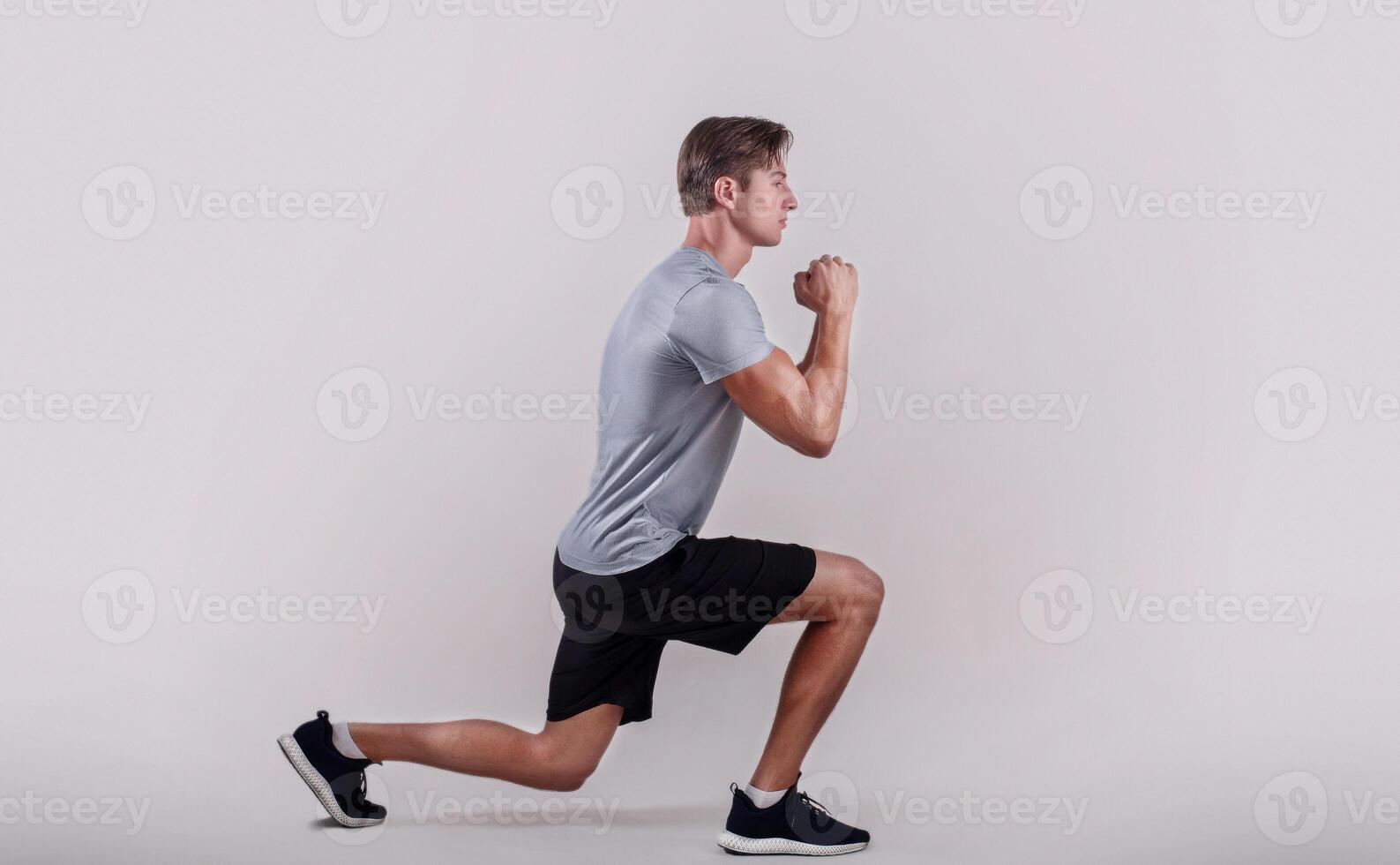 Side view of young fit guy in sportswear doing lunge on light studio background. Professional millennial bodybuilder warming out before strength training, doing aerobic exercises photo