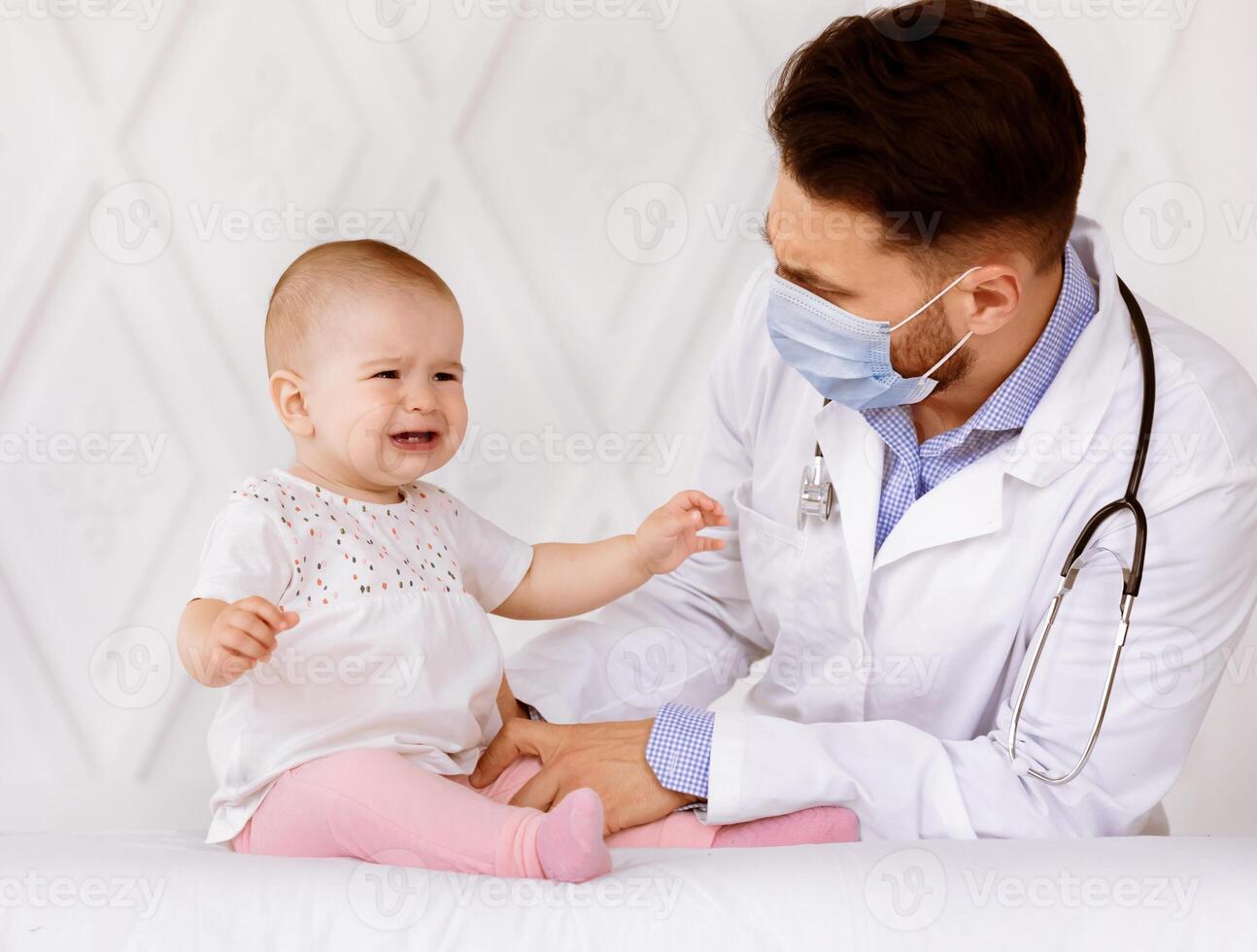 Doctor In Face Mask Examining Little Baby Girl During Child Medical Checkup In Clinic Indoor. Sick Toddler Patient Crying During Appointment In Hospital. Kid's Healthcare Problems Concept photo