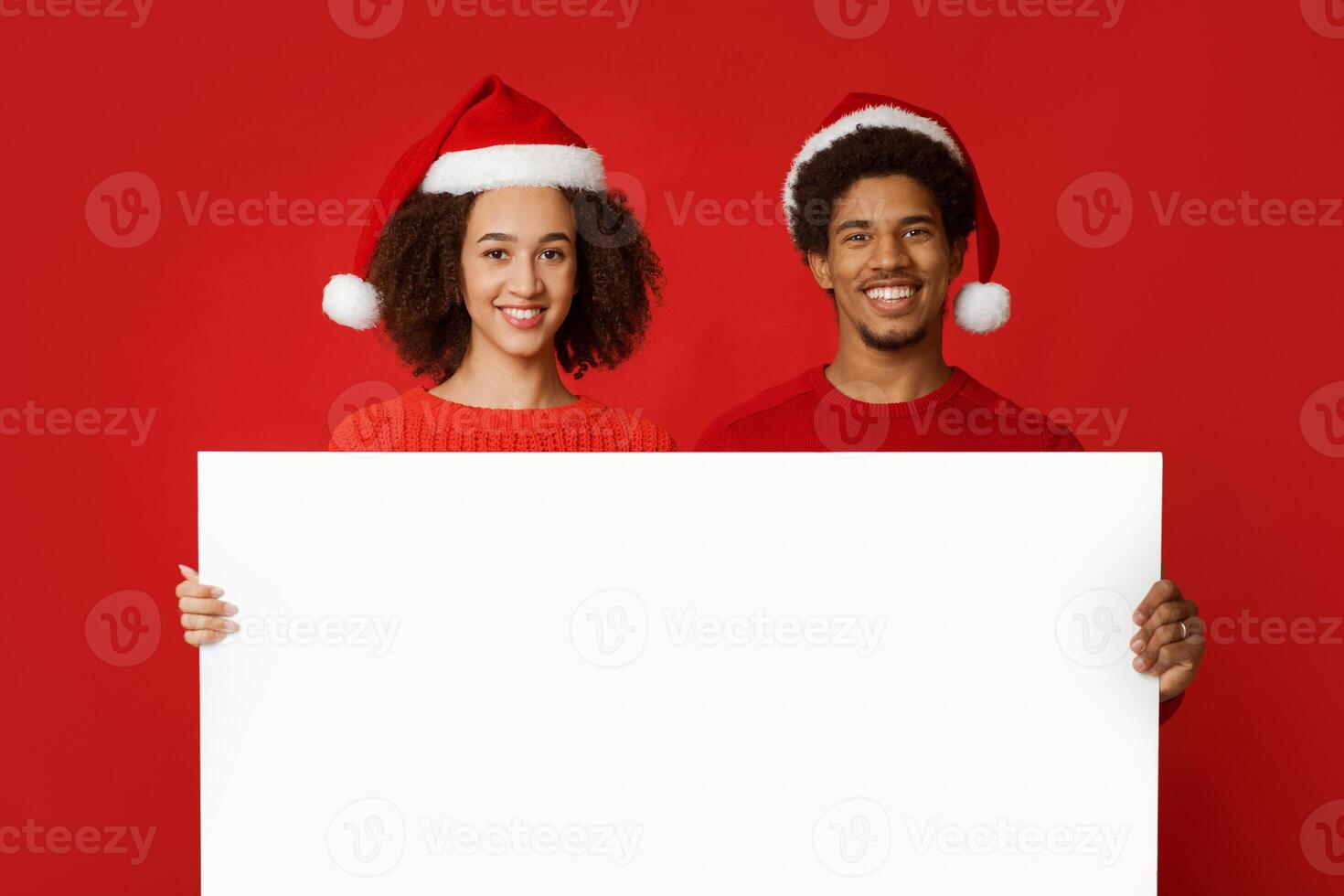Christmas couple holds white board with copy space and advertise New Year season shopping sale concept. Millennial african american happy people in hats, isolated on red background, studio shot photo