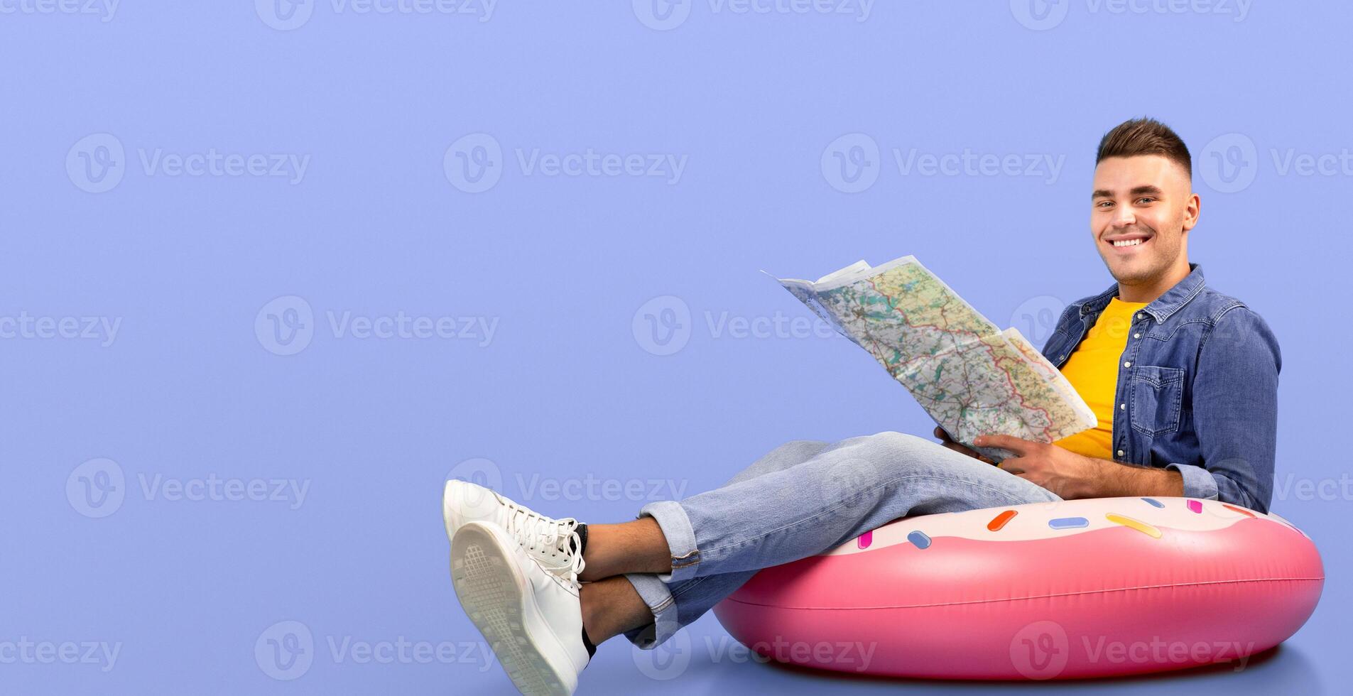 Cool young man sitting on inflatable ring with map, looking at camera and smiling over blue studio background. Full length portrait of tourist imagining his vacation in tropical paradise photo