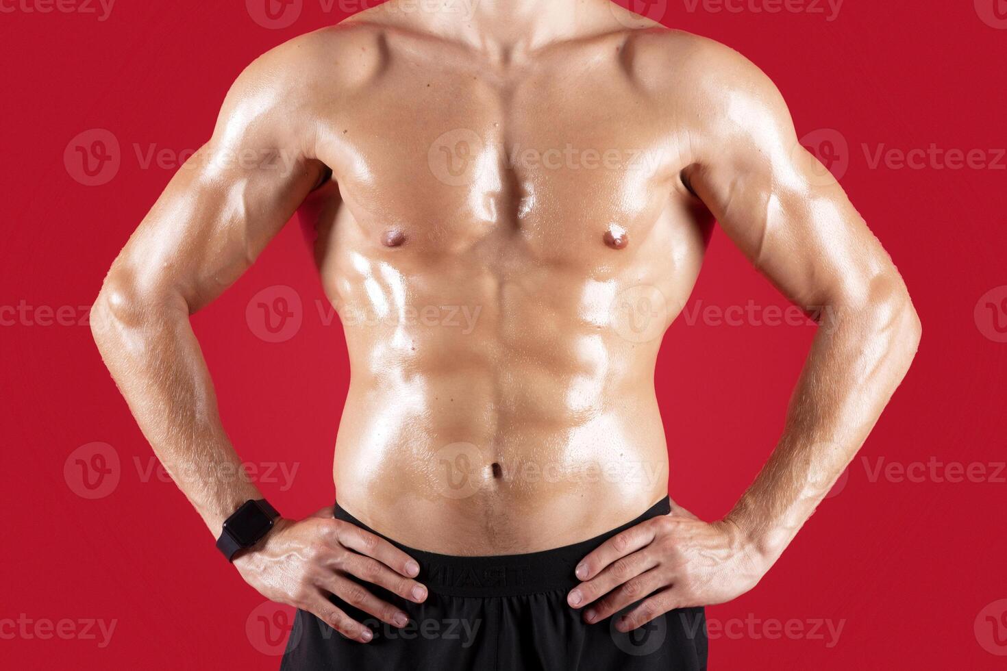 Cropped view of handsome young sportsman with bare torso posing with hands on hips over red studio background. Sexy athletic guy with prominent abs muscles, closeup view. Strength workout concept photo
