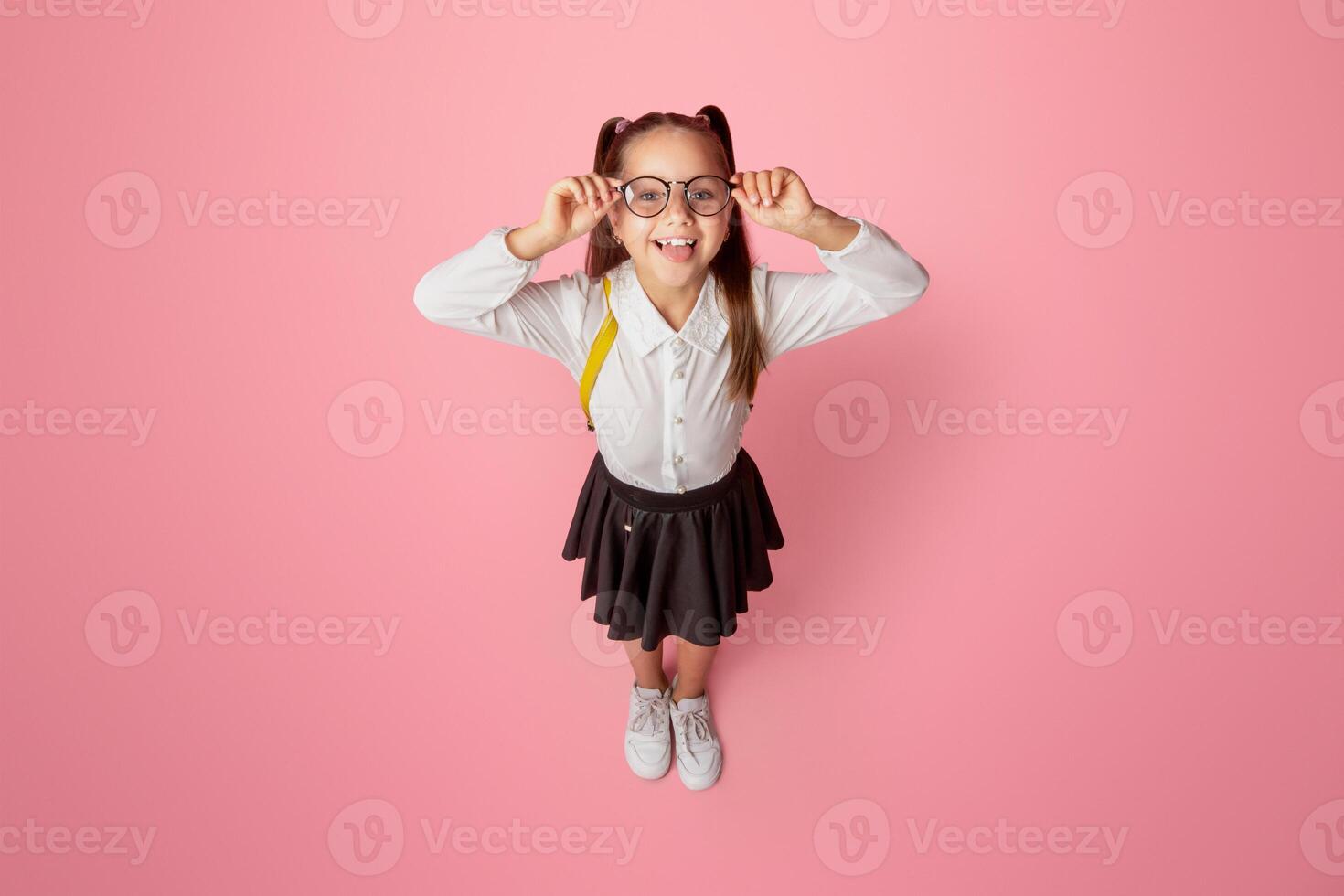 Success in school. Smiling little schoolgirl with glasses and backpack shows tongue and have fun, isolated on pink background, studio shoot photo