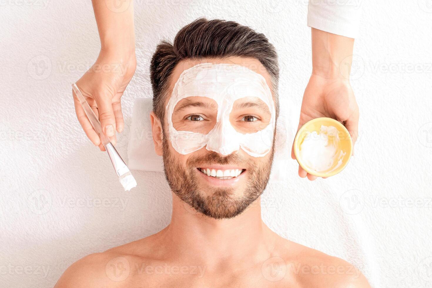 Top view of bearded cheerful man getting face treatment at modern spa salon. Female attendant applying white face mask on middle aged man face. Male face care concept, panorama with free space photo