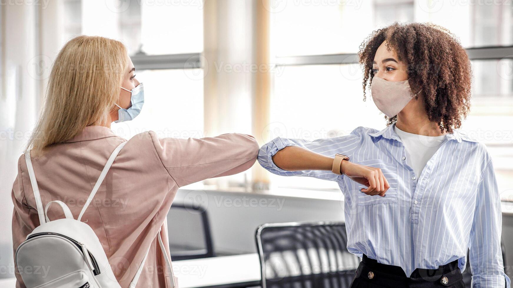 Social distancing and modern greeting during quarantine. African american and european women in protective masks came to work and greet each other with their elbows in interior of coworking office, free space photo