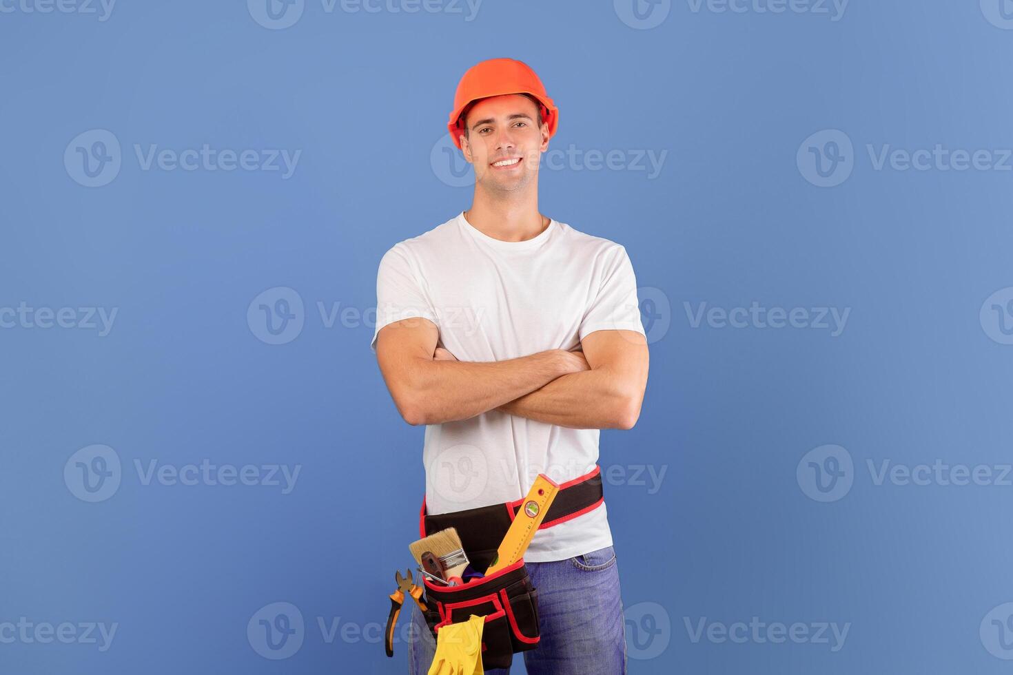 Confident young couple in hard hats ready for house repairment and renovation, millennial spouses in casual clothes standing with folded arms over blue background, man wearing tool belt, copy space photo