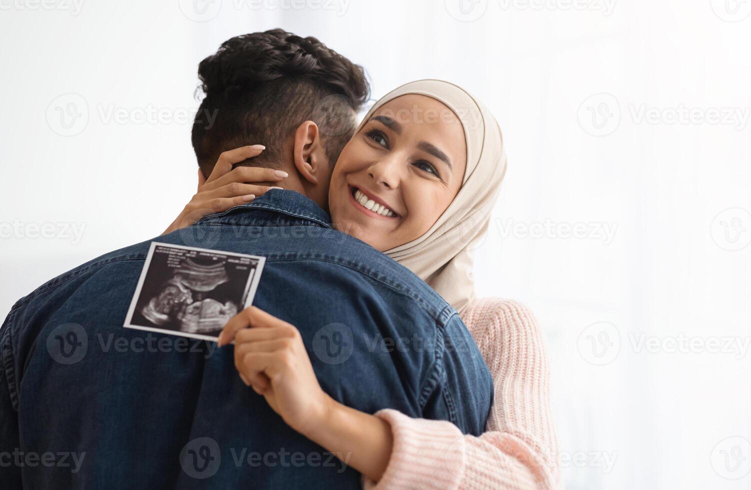 Expecting A Baby. Closeup Portrait Of Cheerful Pregnant Muslim Woman In Hijab Holding Ultrasound Picture And Hugging Her Husband, Standing Near Window At Home, Enjoying Healthy Pregnancy, Copy Space photo