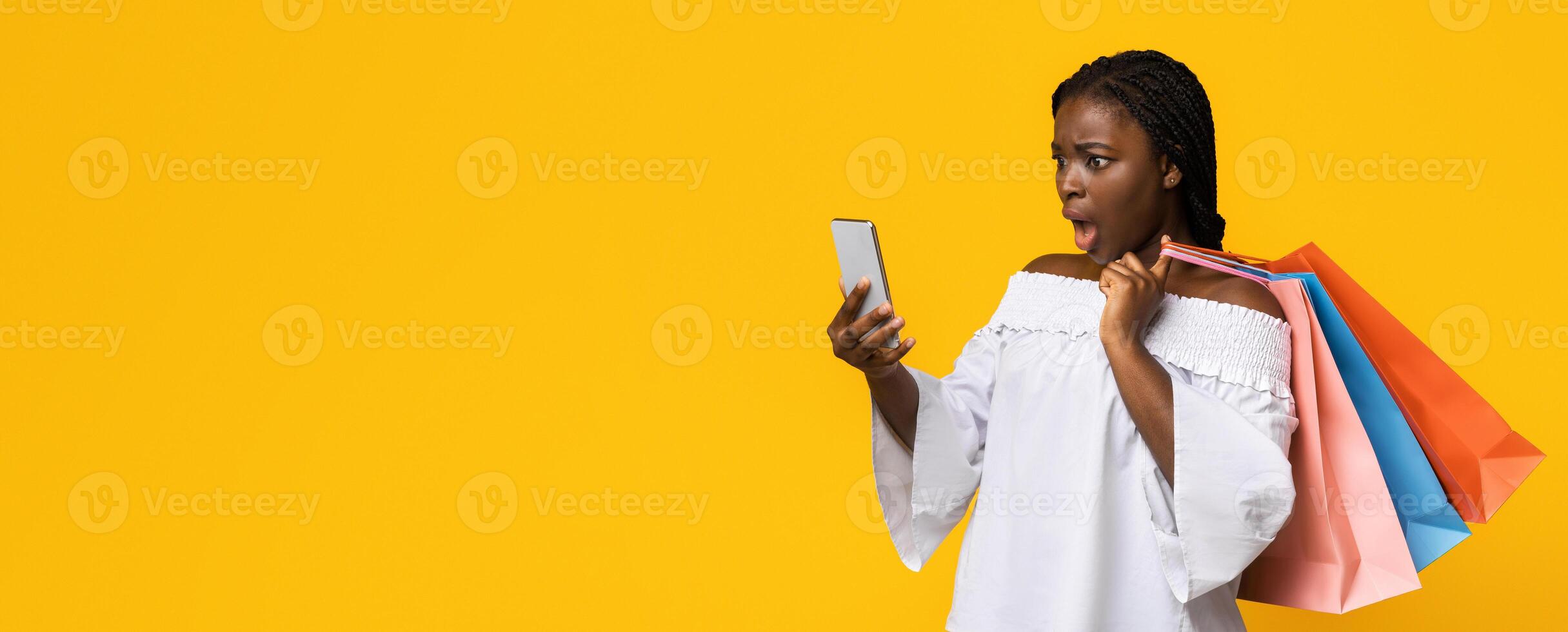 Shocked black shopaholic lady holding smartphone, checking money balance in banking app, carrying lots of shopper bags after big shopping, spent all money, confused woman standing on yellow background photo